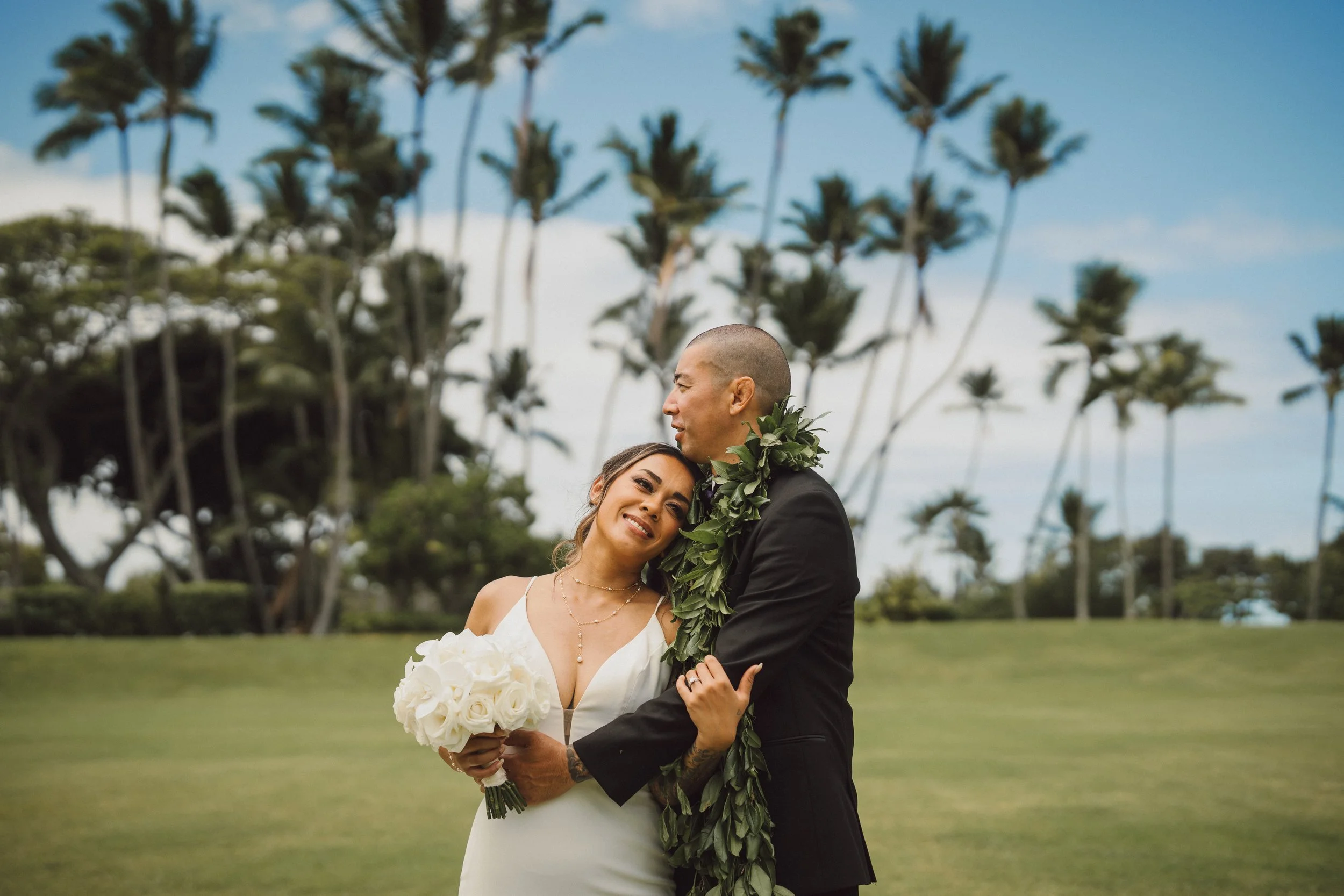 A couple dressed in wedding attire embracing outdoors on a lush green field with tall palm trees in the background. The bride is holding a bouquet of white roses and smiling happily, while the groom, wearing a black suit with a leafy lei, gently lean