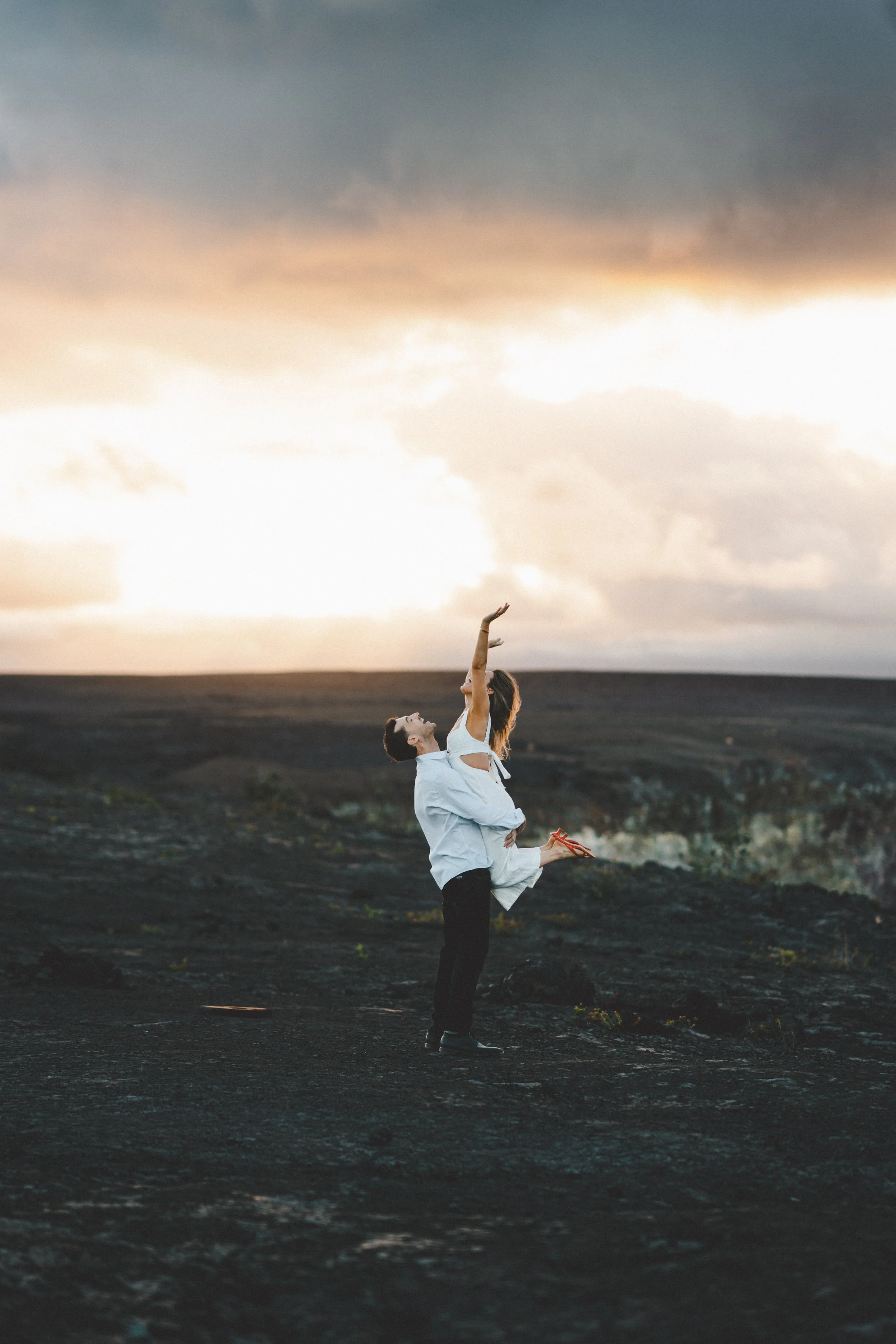 A romantic couple in wedding attire, with the groom lifting the bride in an outdoor landscape at sunset or sunrise, with a cloudy sky.