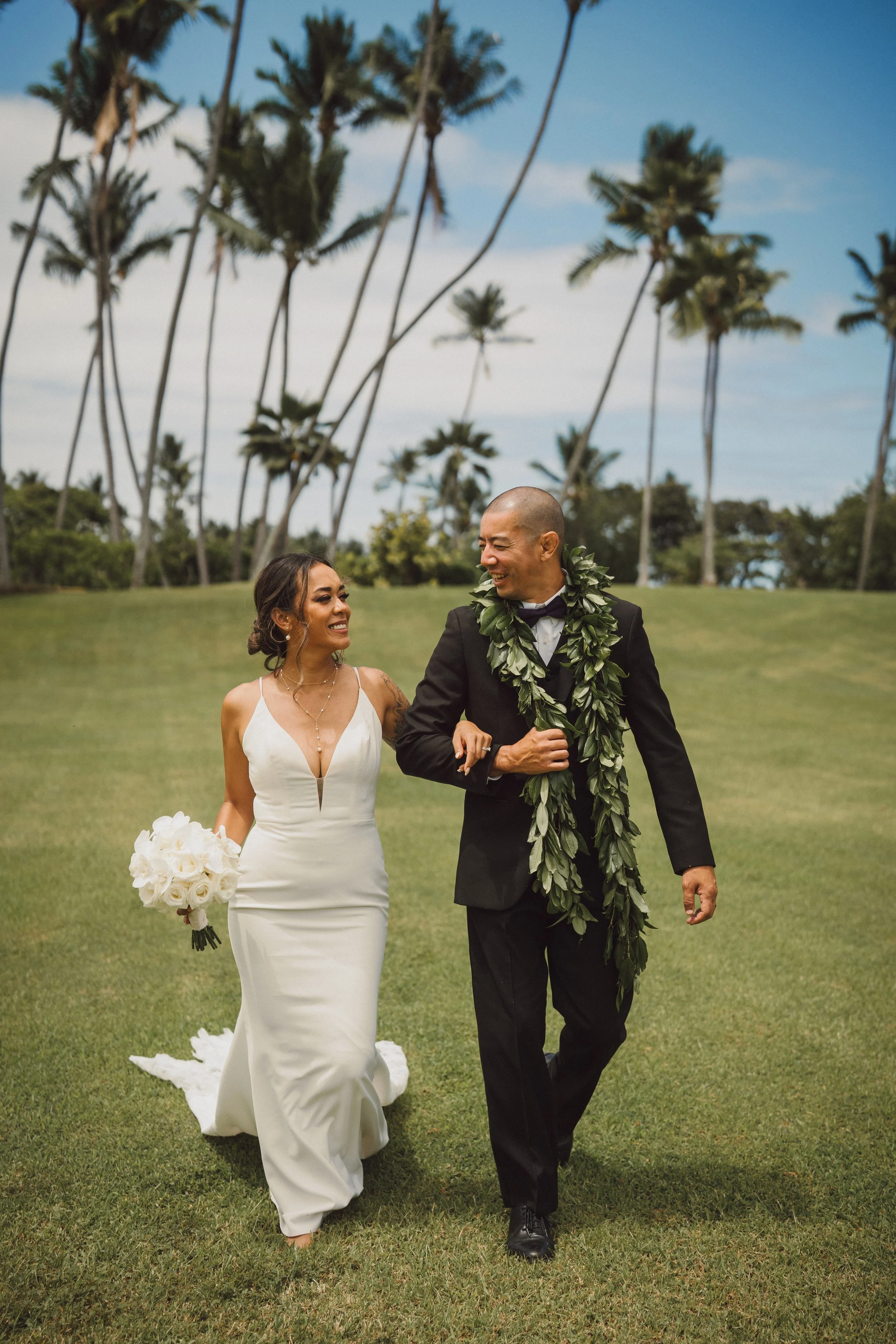 A bride and groom walking together on a grassy field, smiling at each other, with tall palm trees and a blue sky in the background. The bride wears a white sleeveless wedding gown and holds a bouquet of white flowers, while the groom wears a black su
