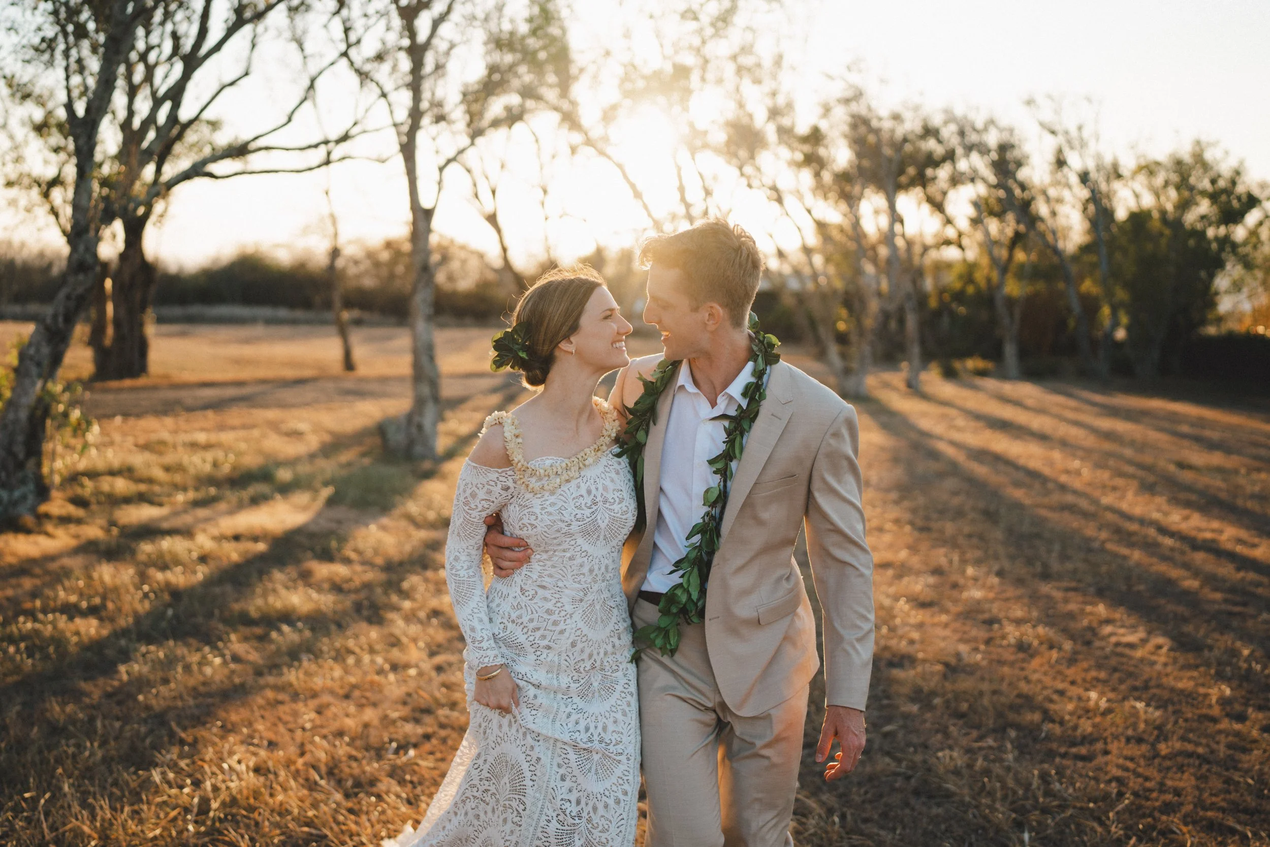 A newlywed couple walking outdoors in a field with trees at sunset, smiling at each other.