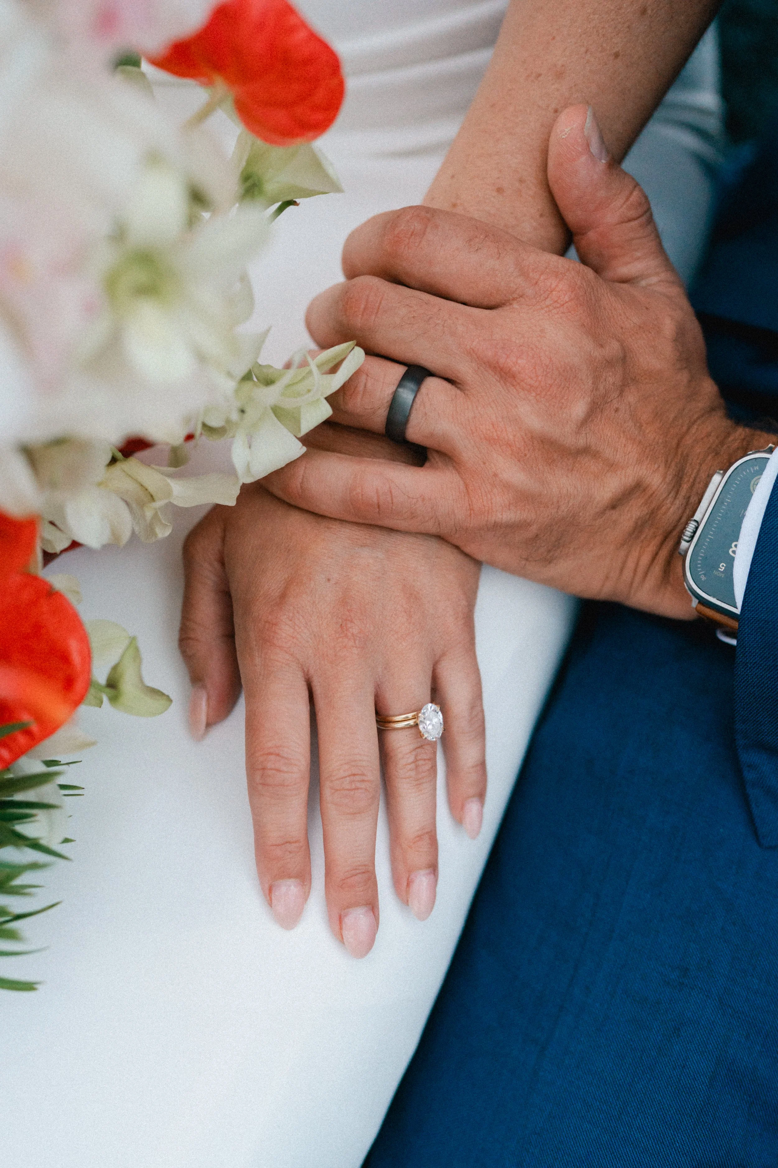 Close-up of newlywed couple's hands showing wedding rings, with flowers and wedding attire visible.