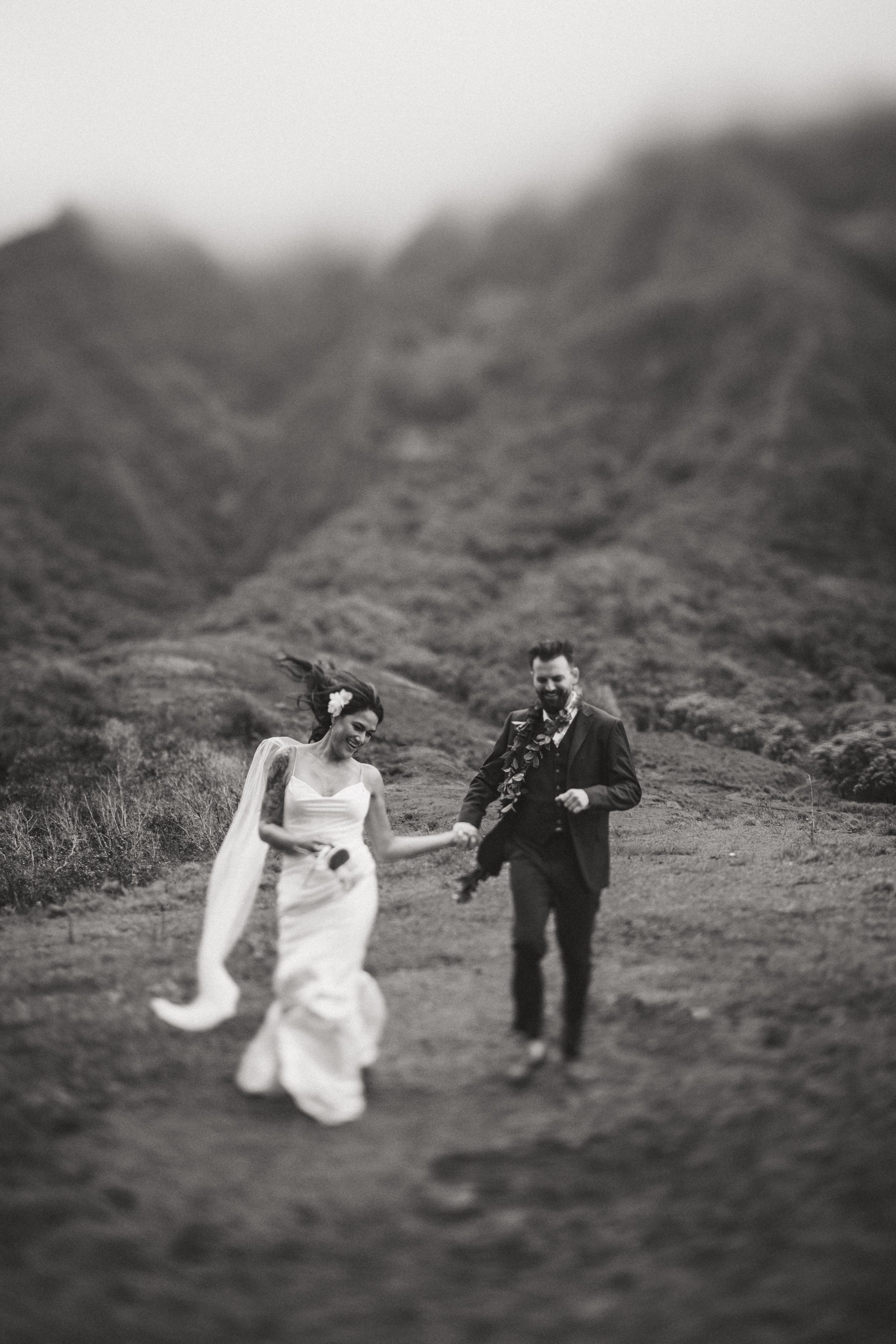 A black and white photo of a bride and groom walking hand in hand outdoors, with mountains in the background.