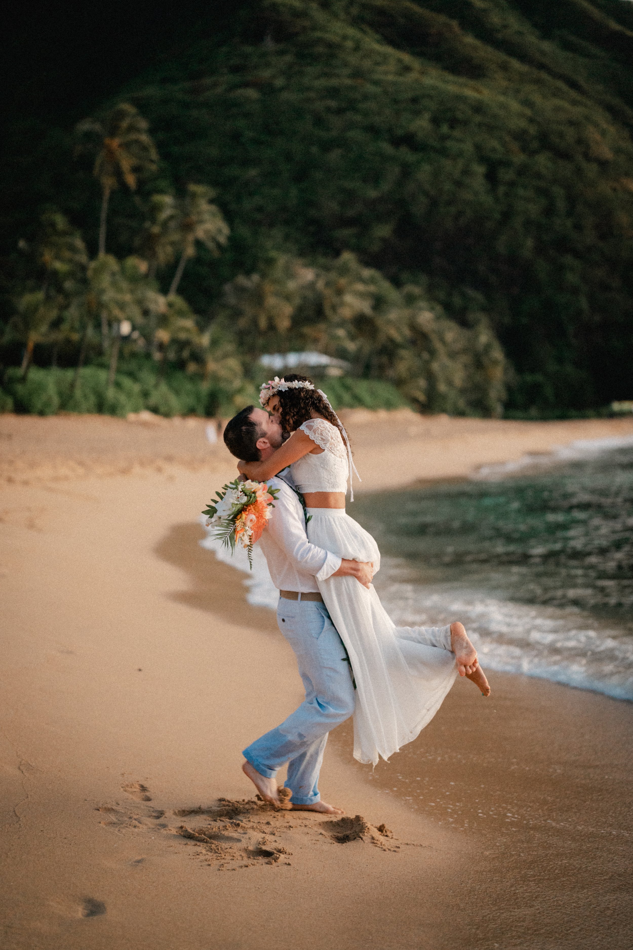 A couple in wedding attire embraces and kisses on the beach, with greenery and hills in the background.