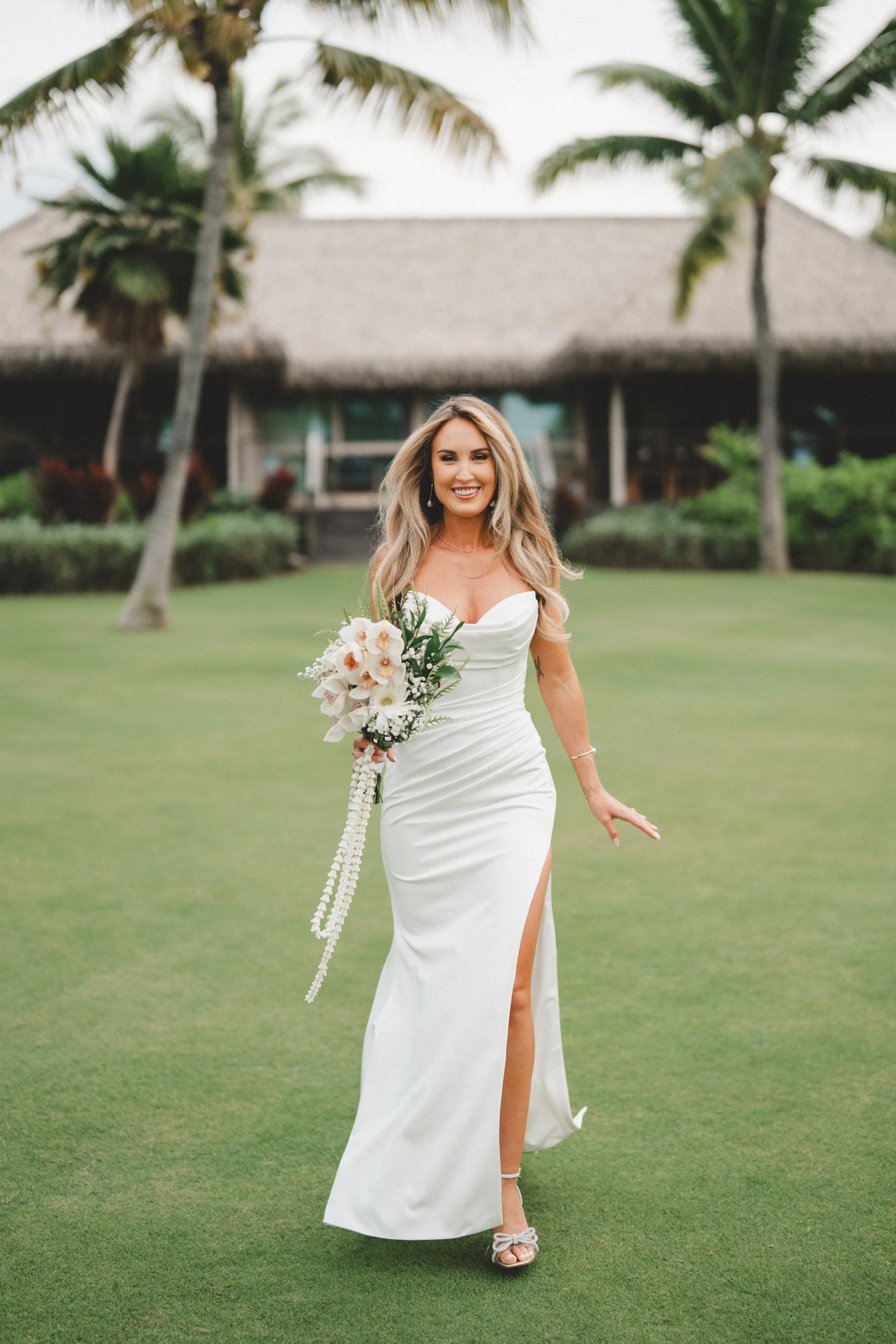 A woman in a white wedding dress holding a bouquet of flowers, walking on a grassy area with palm trees and a building in the background.