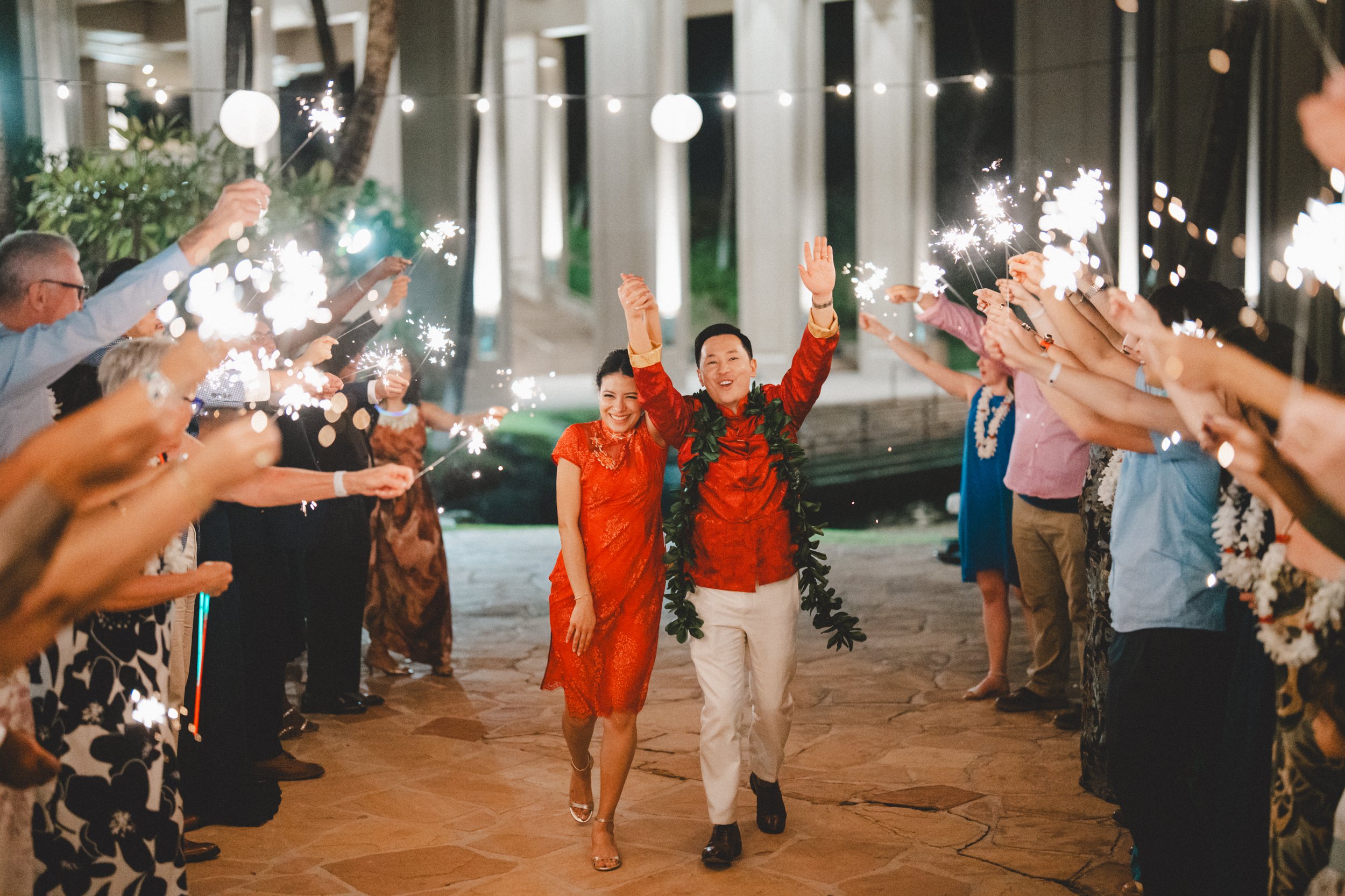 A couple dressed in red walking through a celebration with sparklers, people forming a corridor on both sides, in an outdoor area decorated with string lights at night.