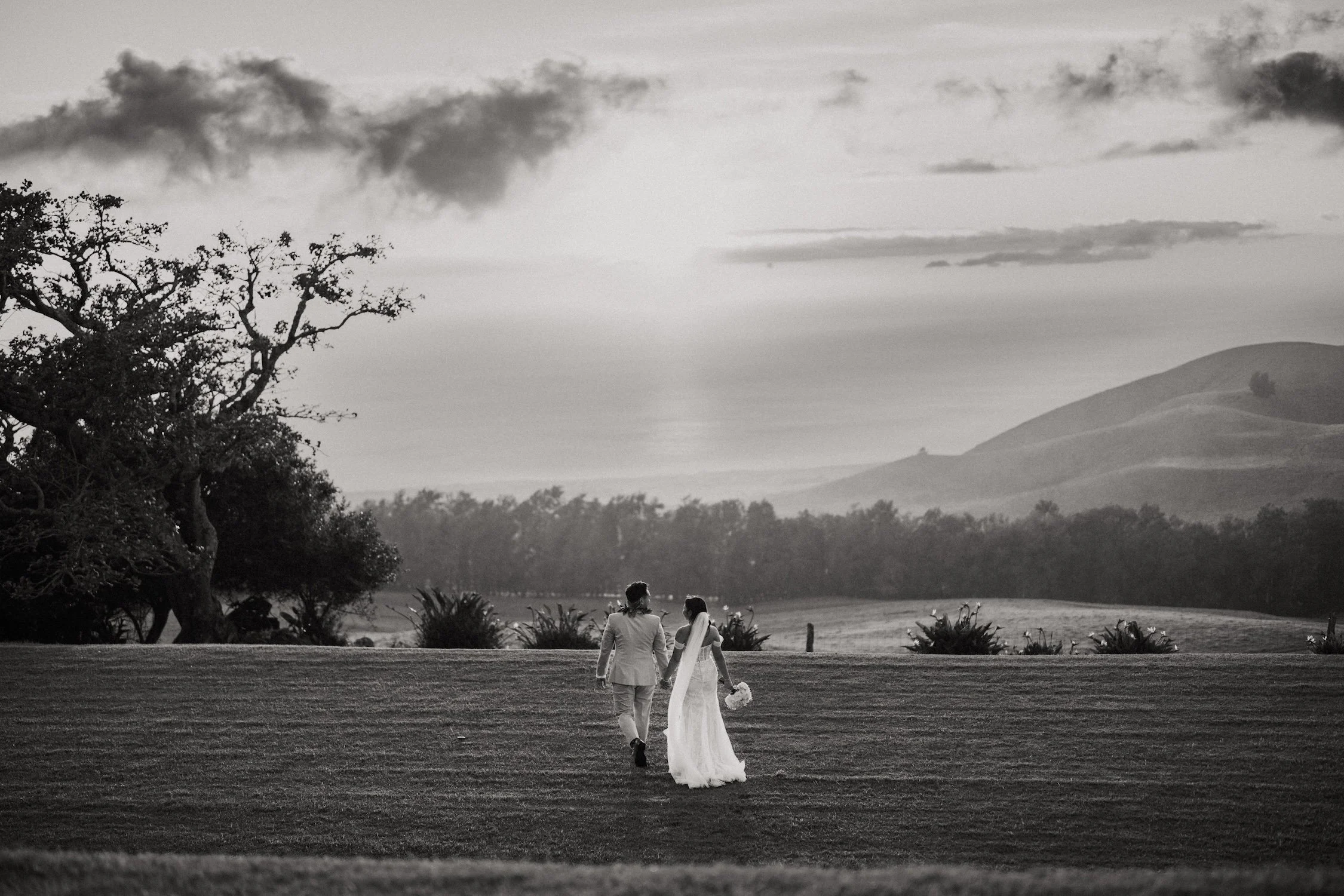 A black and white photo of a bride and groom walking hand in hand across a grassy field, with trees and hills in the background.