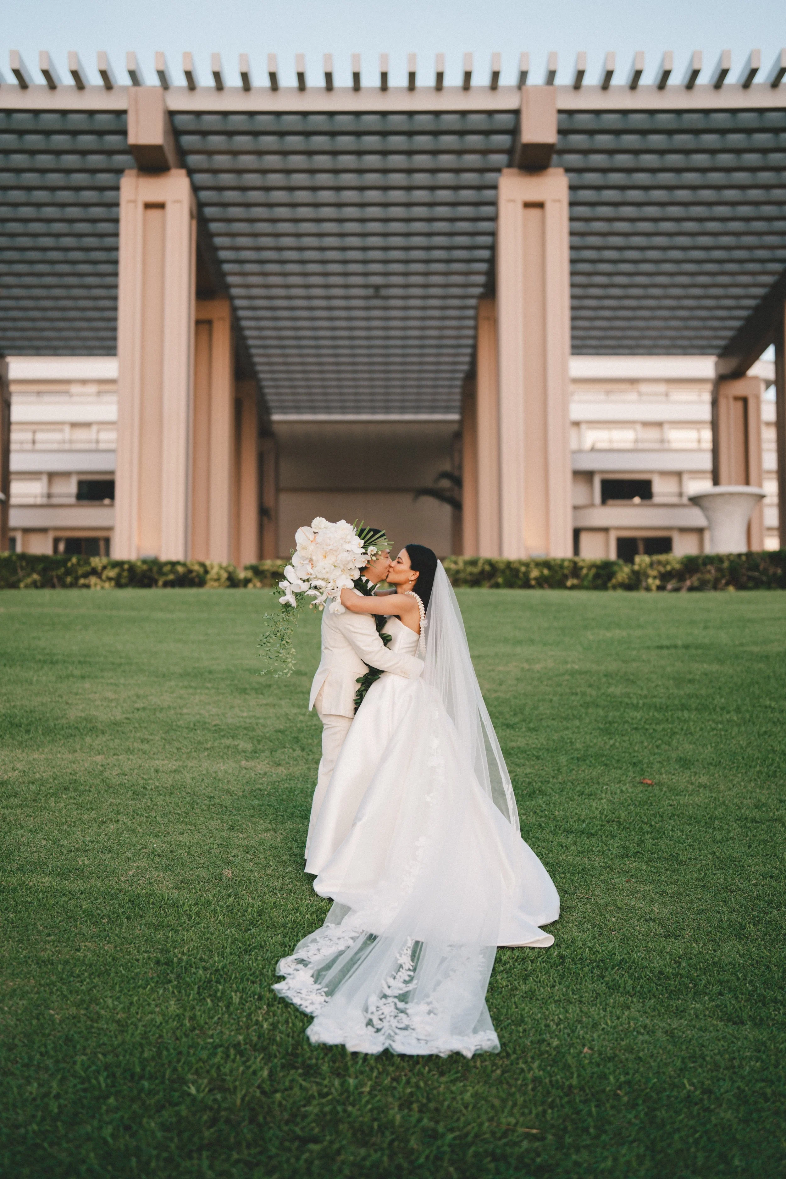 Bride and groom kissing outdoors on a grassy area with a modern architectural building in the background.
