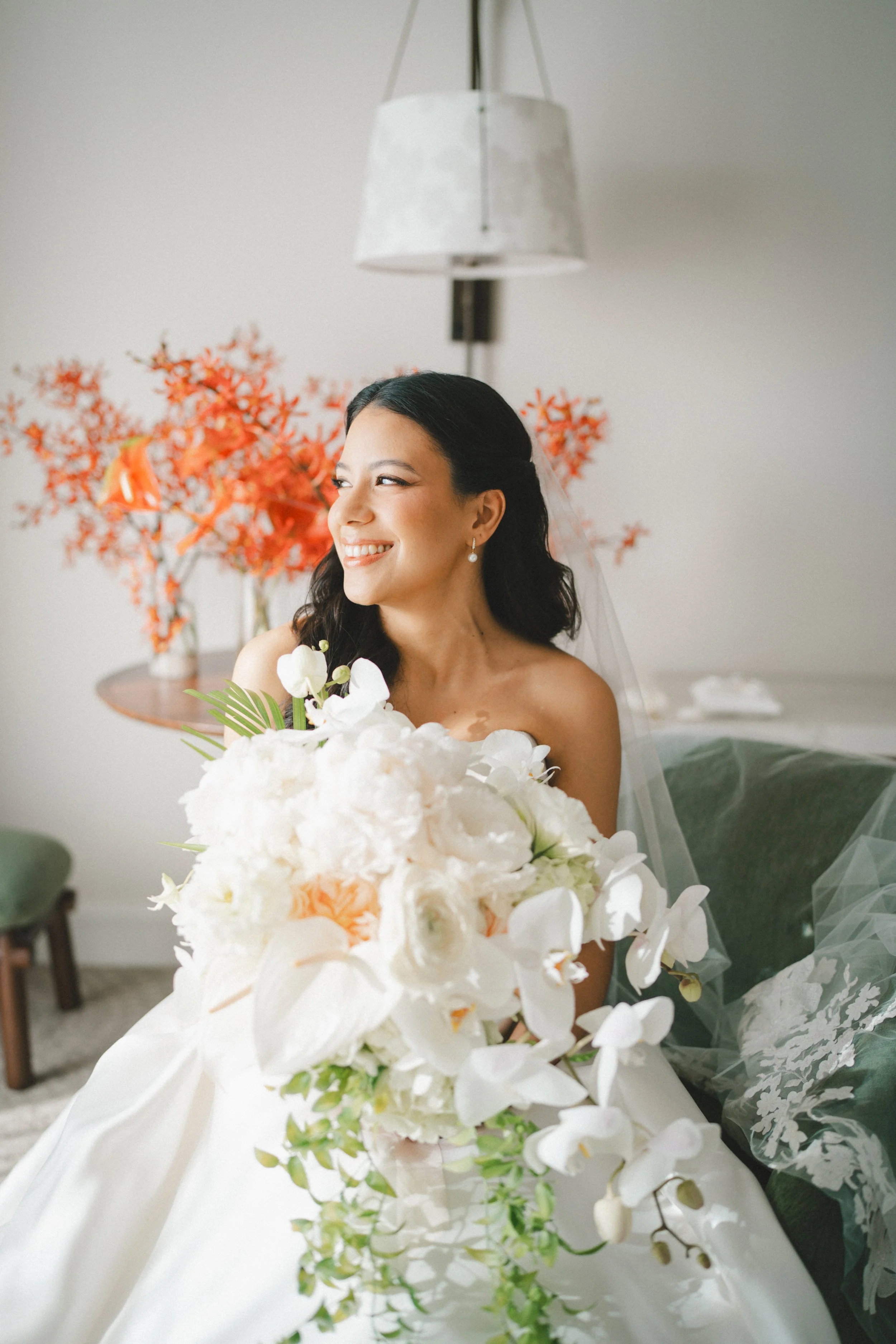 A smiling bride with dark hair, wearing pearl earrings and a veil, holding a large bouquet of white flowers, seated indoors with orange flowers and a lamp in the background.