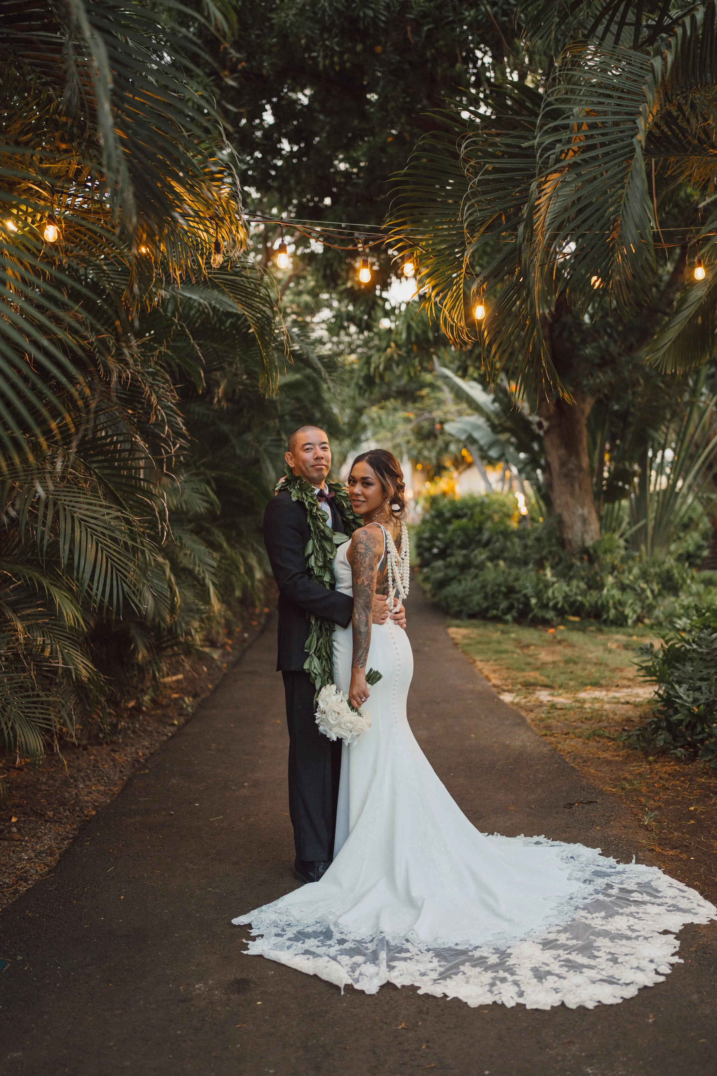 Bride and groom embracing on a garden pathway under string lights, surrounded by lush tropical plants during sunset.