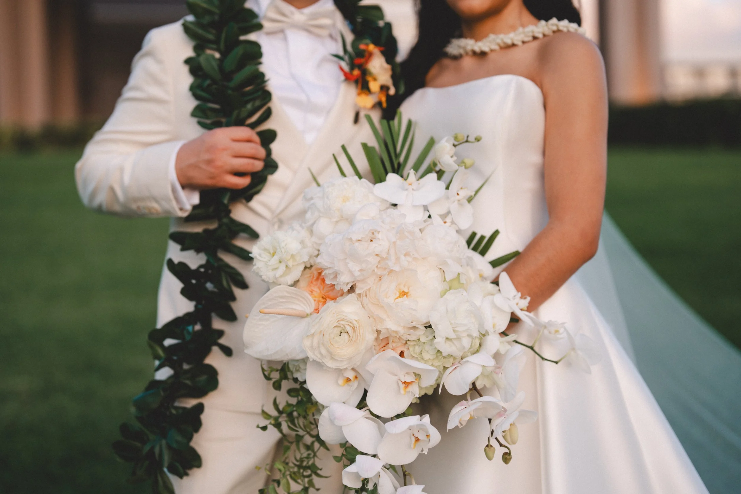 Bride and groom in wedding attire, holding a large white wedding bouquet with orchids and greenery, outdoors during sunset.