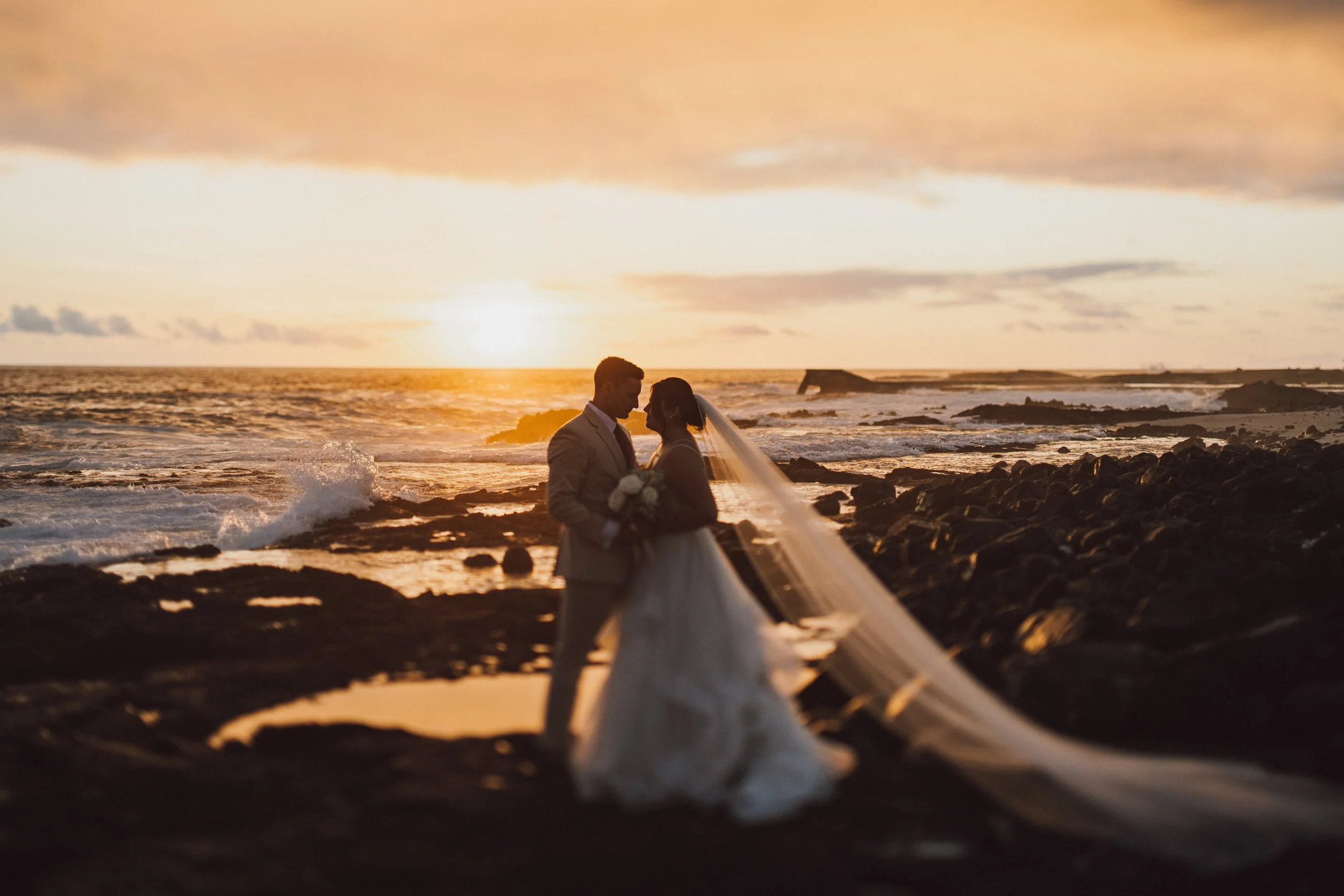 Silhouetted bride and groom standing on rocky beach at sunset, facing each other, with ocean waves crashing behind them.