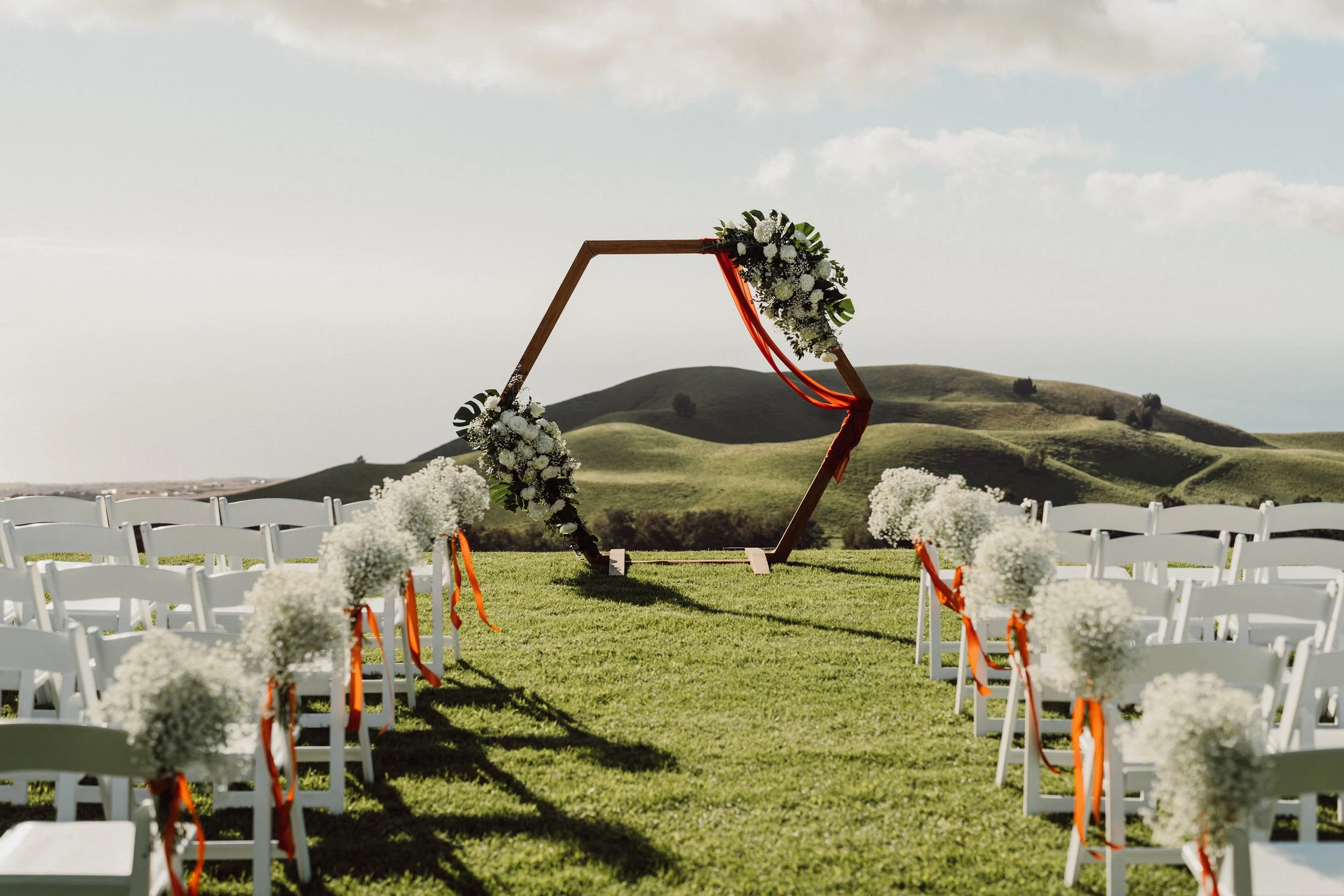 Outdoor wedding ceremony setup with white chairs decorated with flowers and ribbons, a geometric wooden arch with floral arrangements, green rolling hills in the background, and a partly cloudy sky.