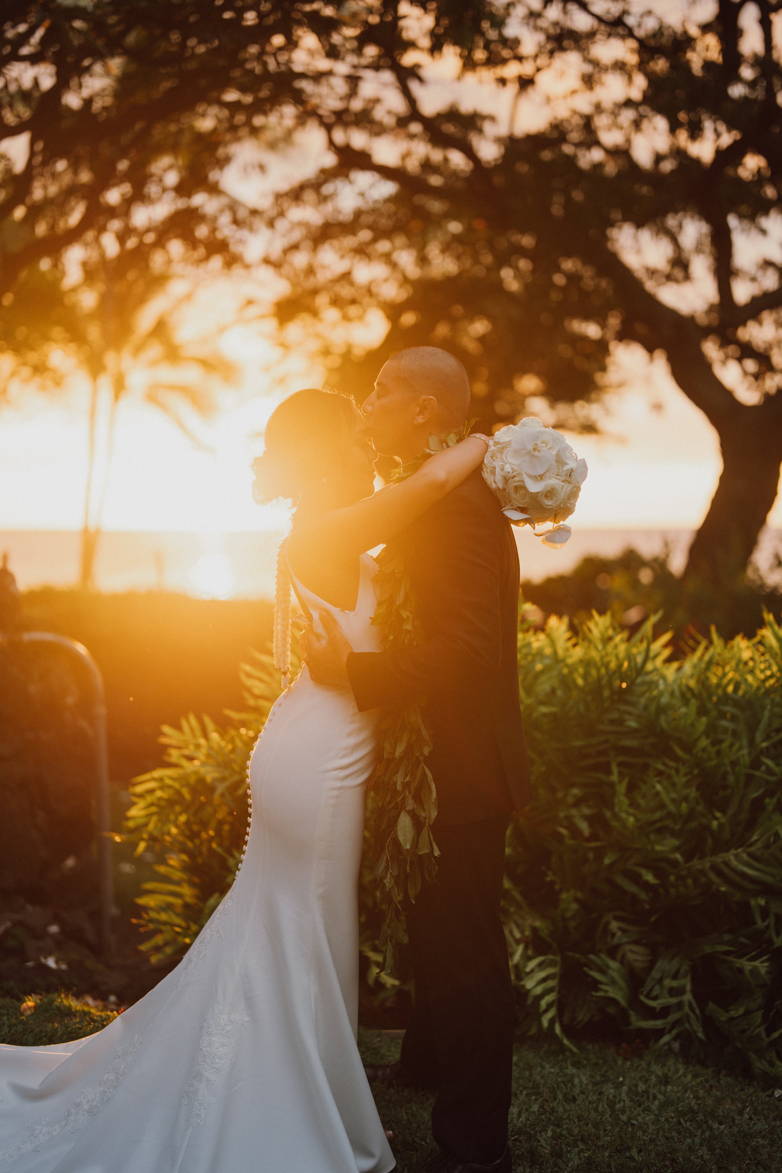 A bride and groom embrace during their wedding at sunset with ocean and trees in the background.