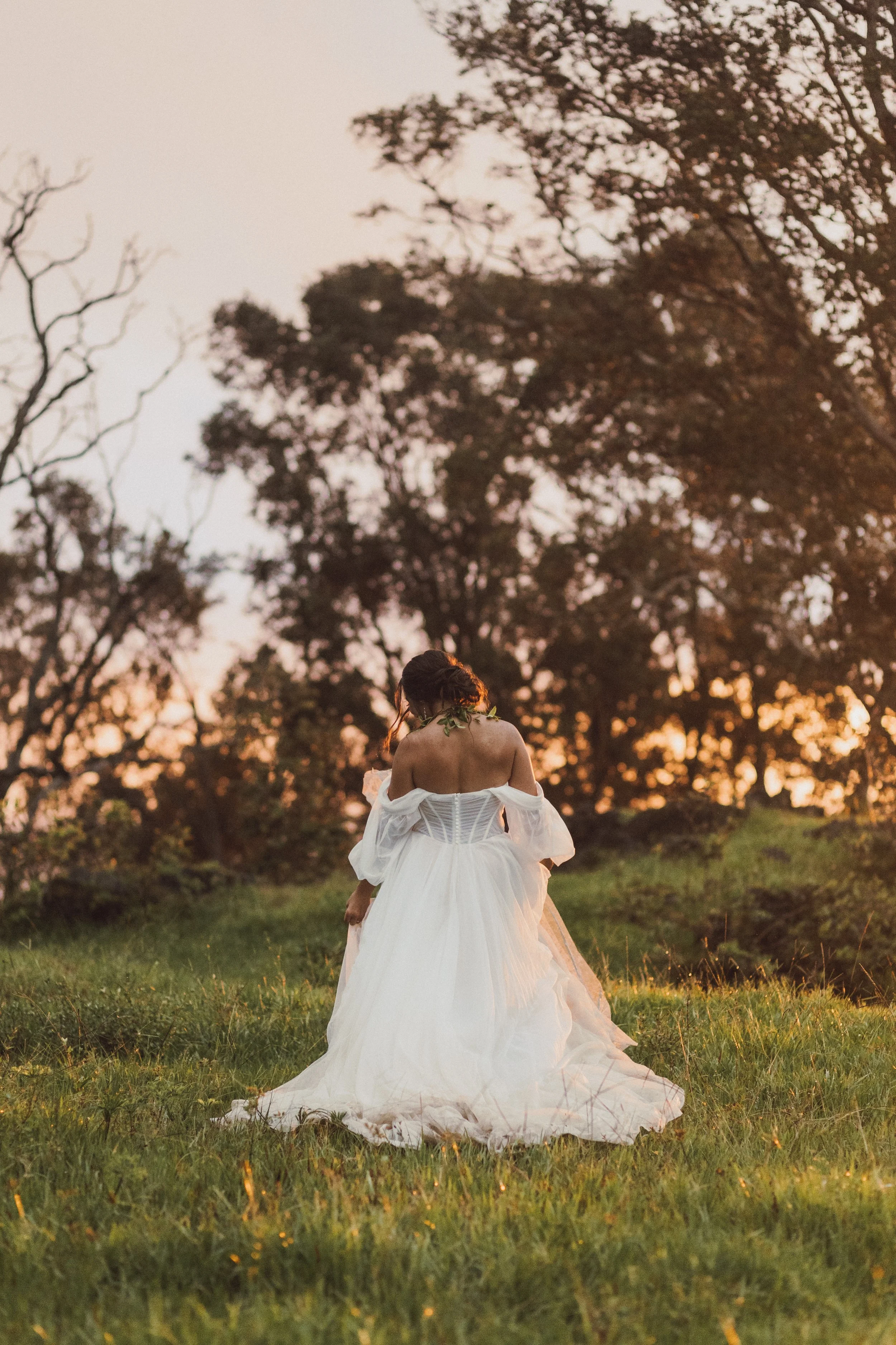A woman in a white wedding dress standing outdoors on grass during sunset, back facing the camera, with trees in the background.