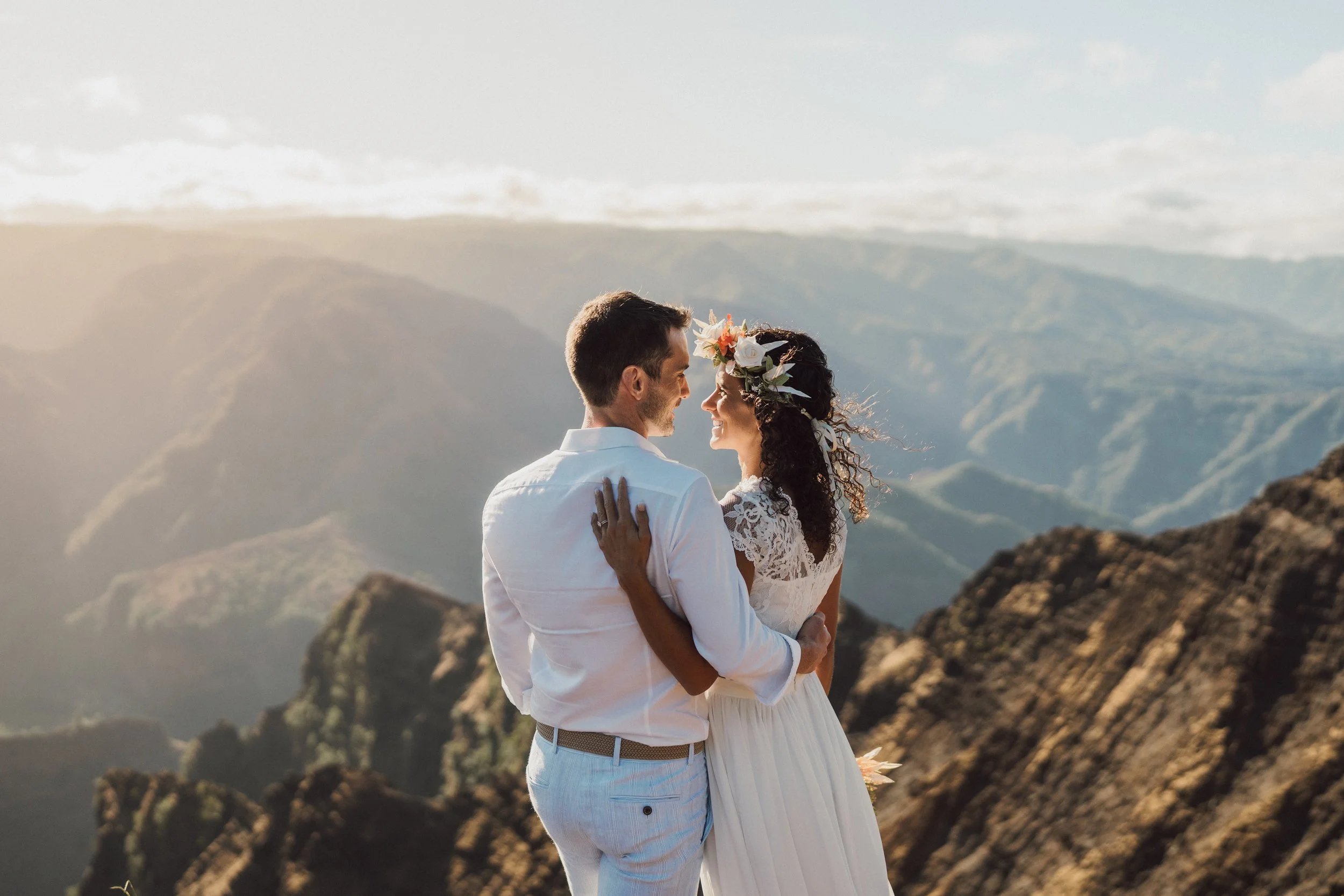 A newlywed couple in wedding attire embracing on a mountain with a scenic view of rolling hills and mountains at sunset or sunrise.