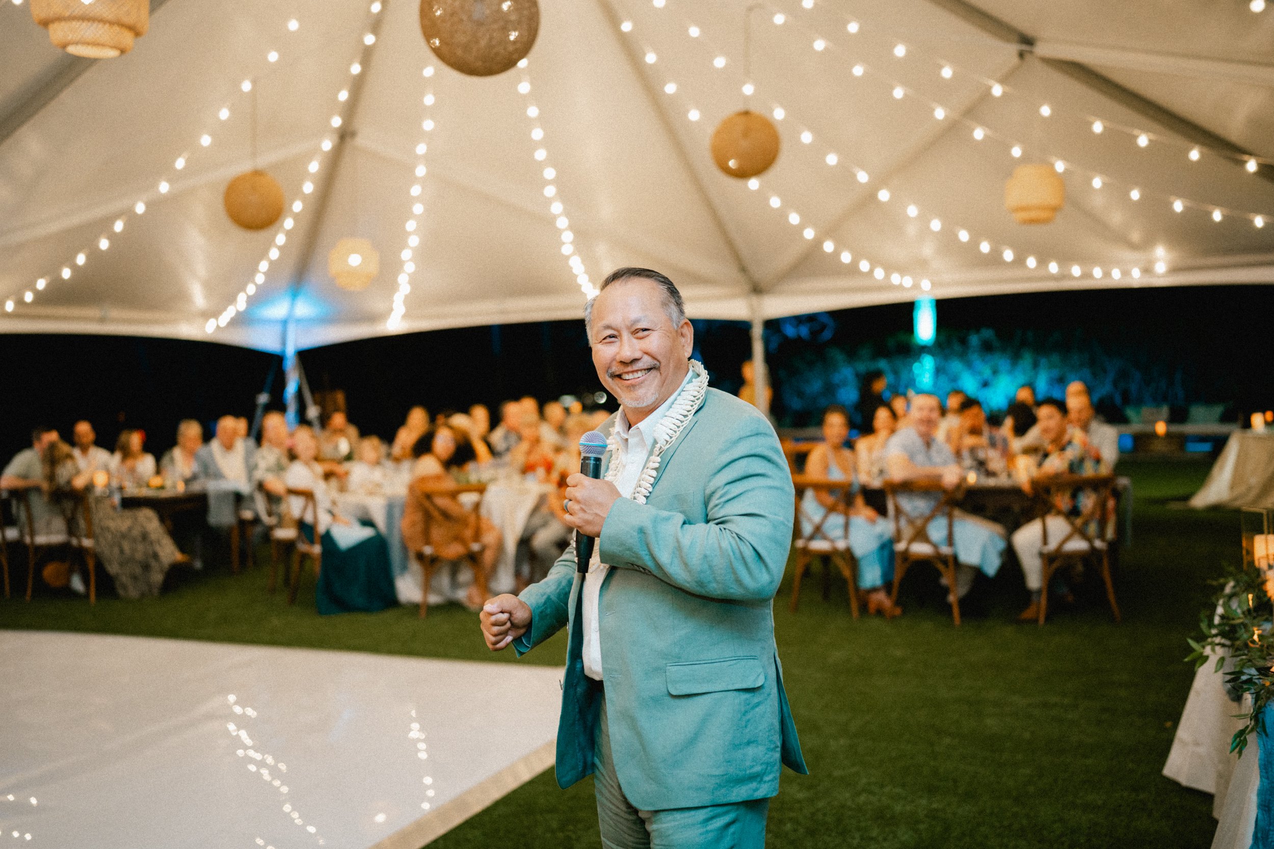 A man in a light blue suit and white shirt smiling while holding a microphone at an outdoor event under a large tent with string lights.