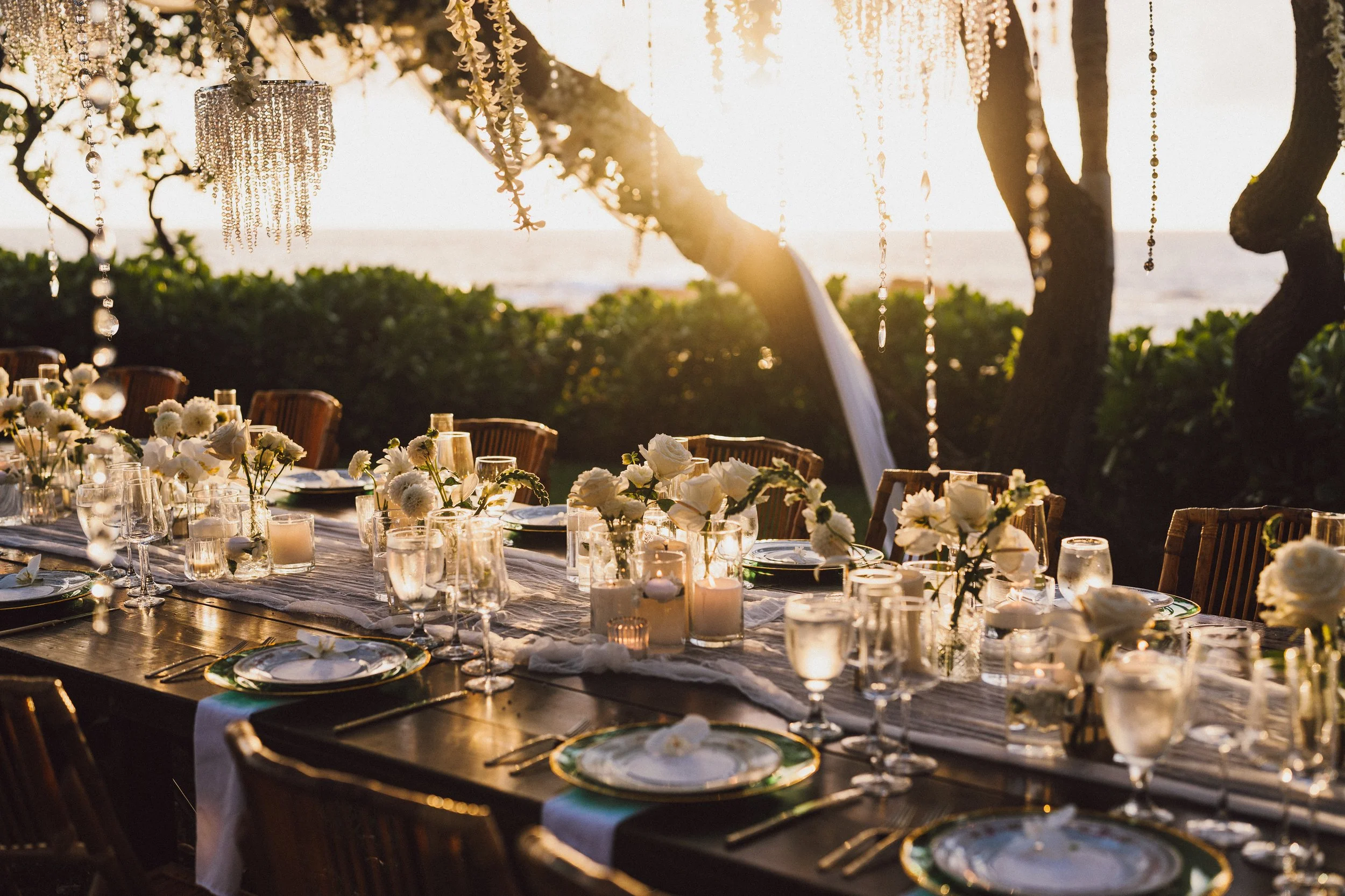 Elegant outdoor dining table set with white flowers in vases, candles, and glassware, beneath hanging crystal chandeliers, with trees and a sunset in the background.