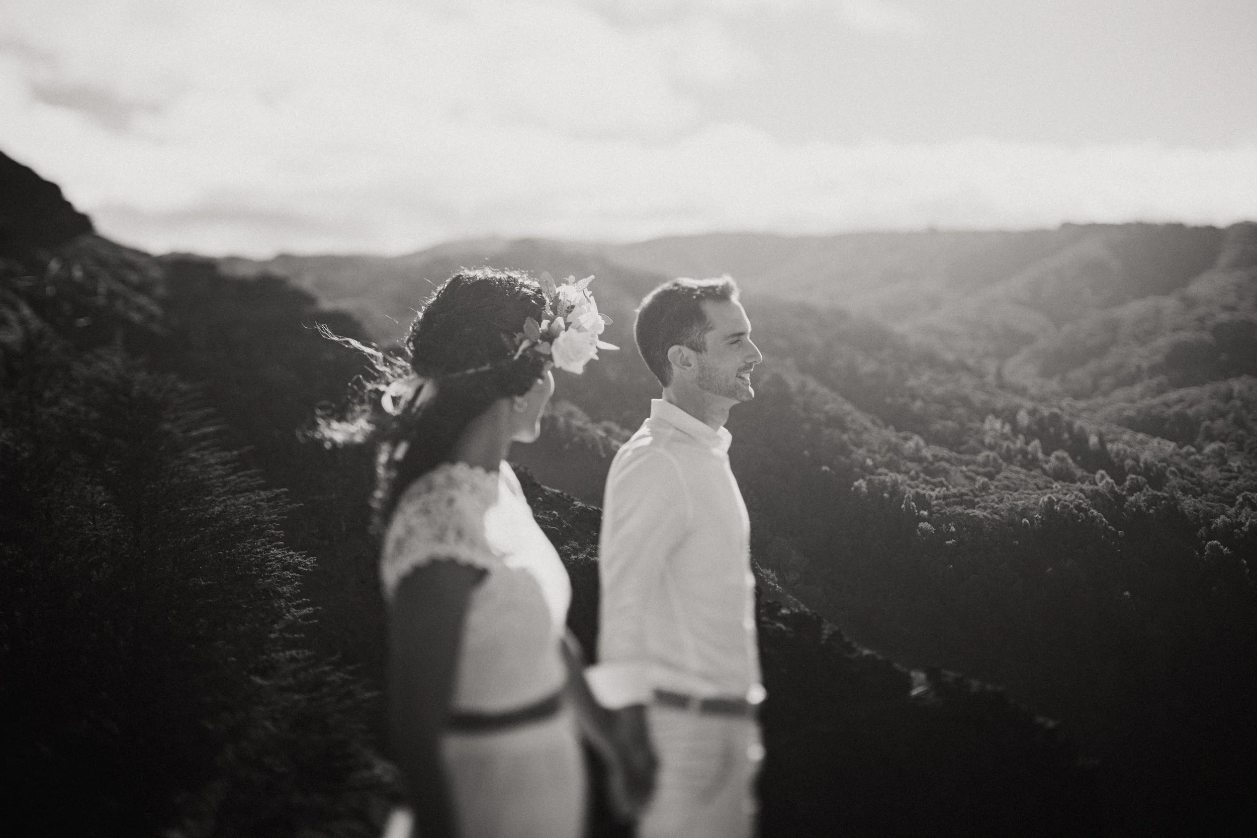 Black and white photo of a couple standing outdoors on a mountain with a landscape of rolling hills in the background. The woman has curly hair with a flower crown, and the man is smiling, both facing to the right.