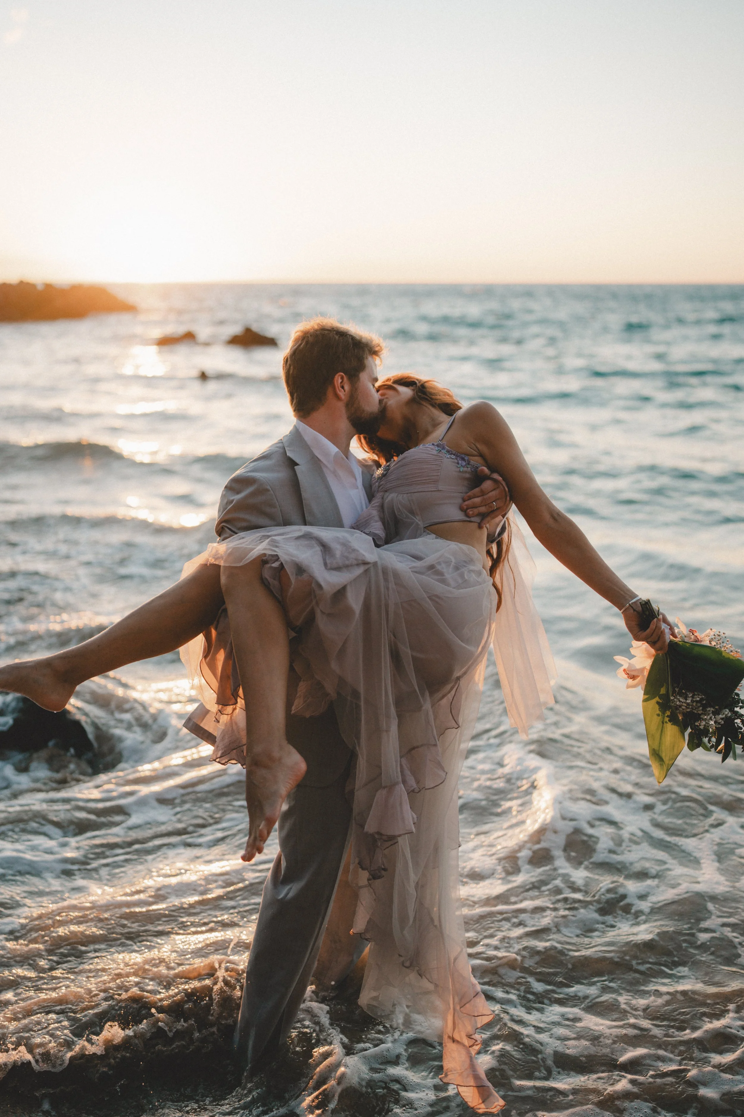 A couple kissing on the beach at sunset, the man in a suit carrying the woman in a flowing dress, holding a bouquet of flowers.