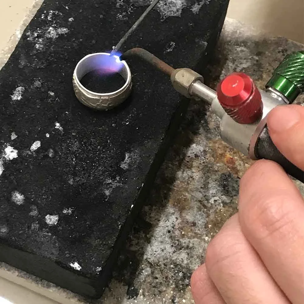 Close-up of a person using a handheld torch to weld or heat a metal ring on a work surface with black and burnt marks.