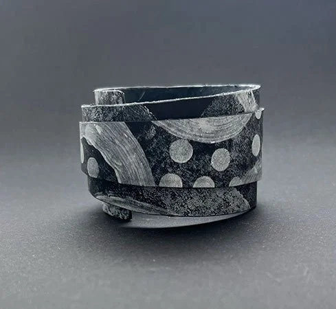 Close-up of a black and gray clay or ceramic ring with abstract patterns and dots, on a dark surface with a gray background.