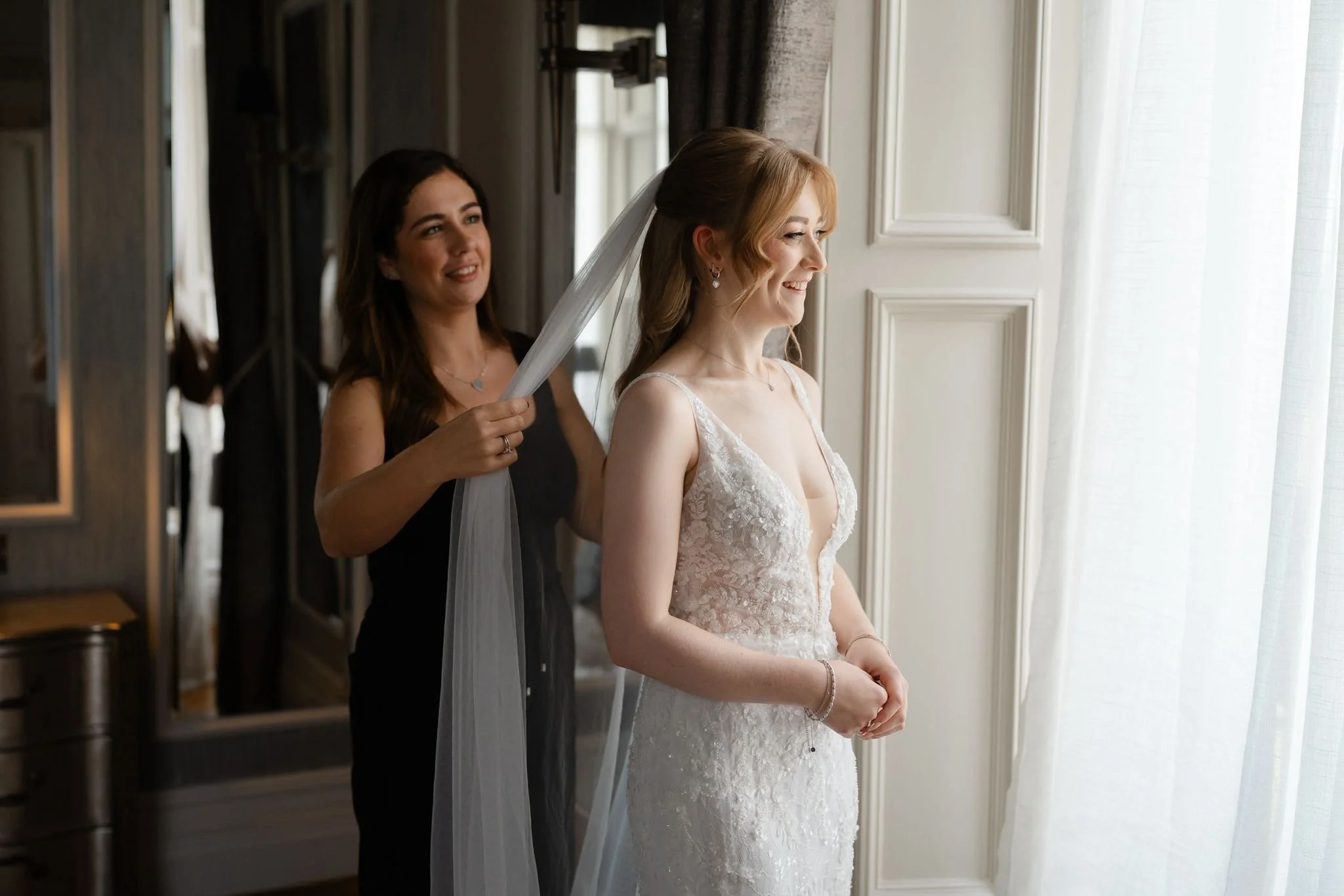 A woman with wavy hair and dressed in an off-shoulder dress is smiling while a person adjusts her hair and adds a headpiece.