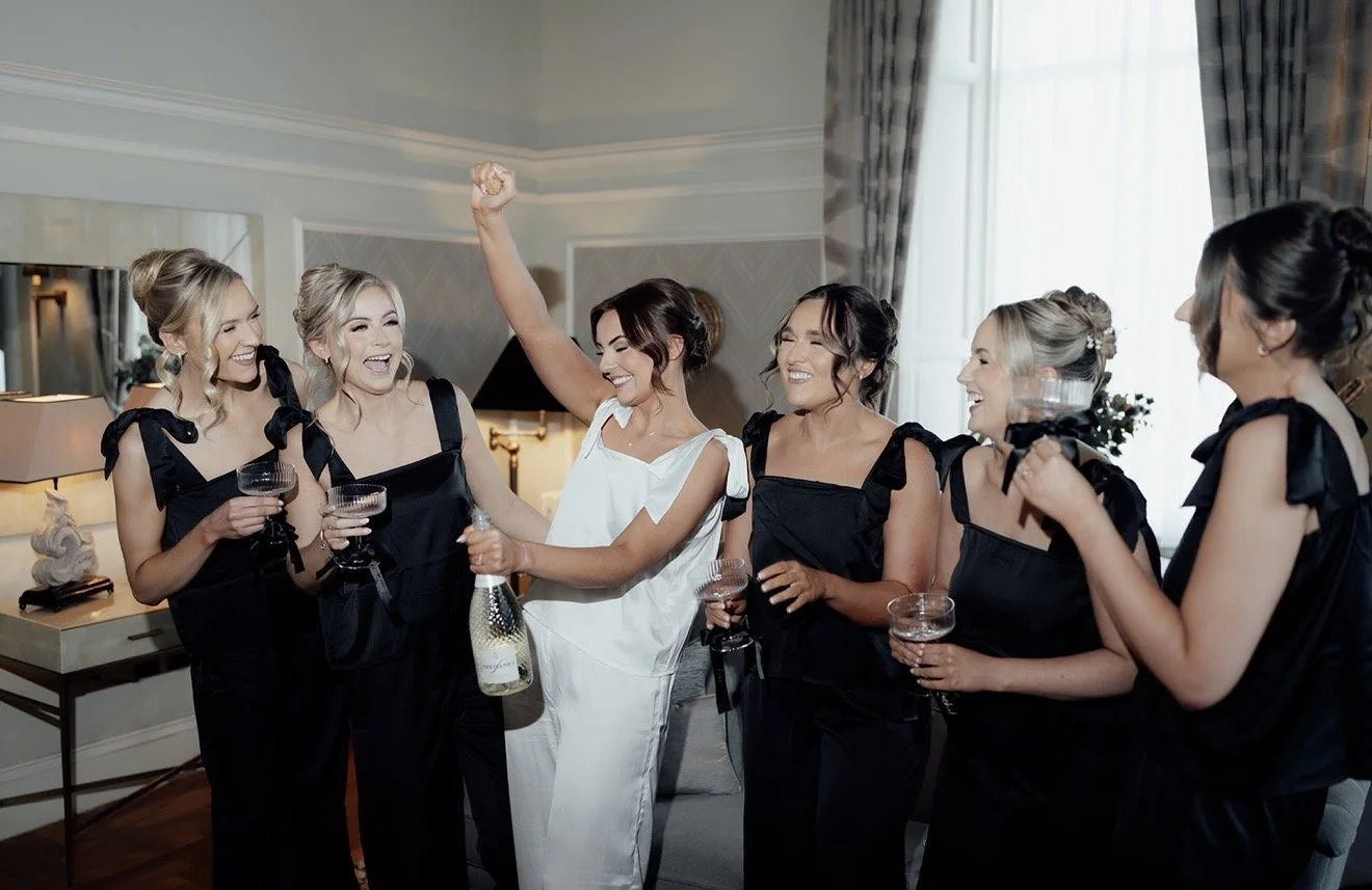 Six women, including a bride in a white gown and five bridesmaids, standing outdoors with trees in the background, holding bouquets and smiling.