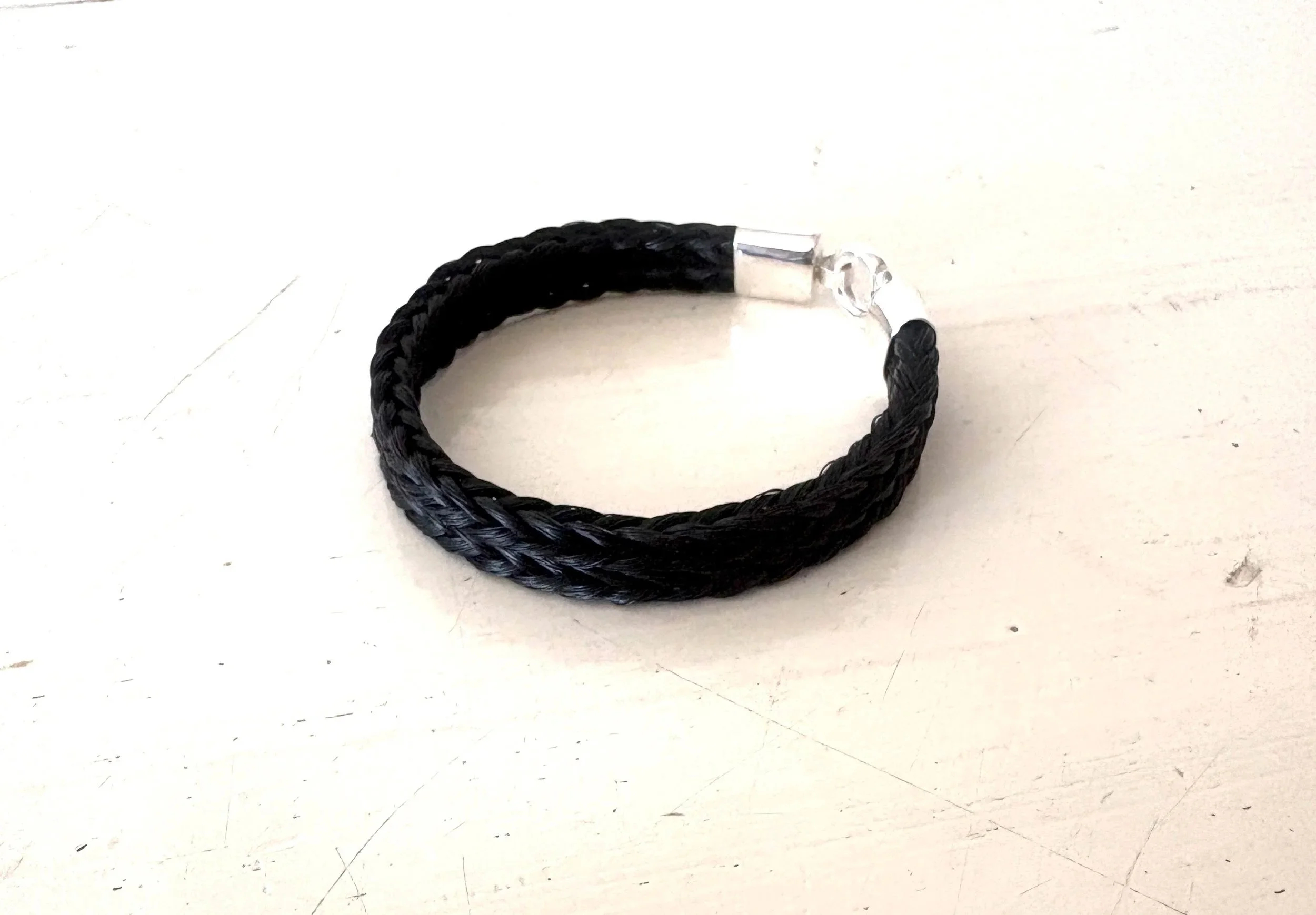 Kraftigt armbånd syet sammen af to firkantede flet. Afsluttet med ovale håndsmedede sølvender og cirkellås i sølv. Forbrug 3-4 blyanttykkelser halehår- 50cm. længde. Pris 2400,- forgyldt sølv plus 200,-