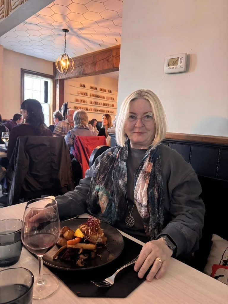 A woman sitting at a restaurant table with a glass of red wine and a plated meal, with other diners visible in the background.
