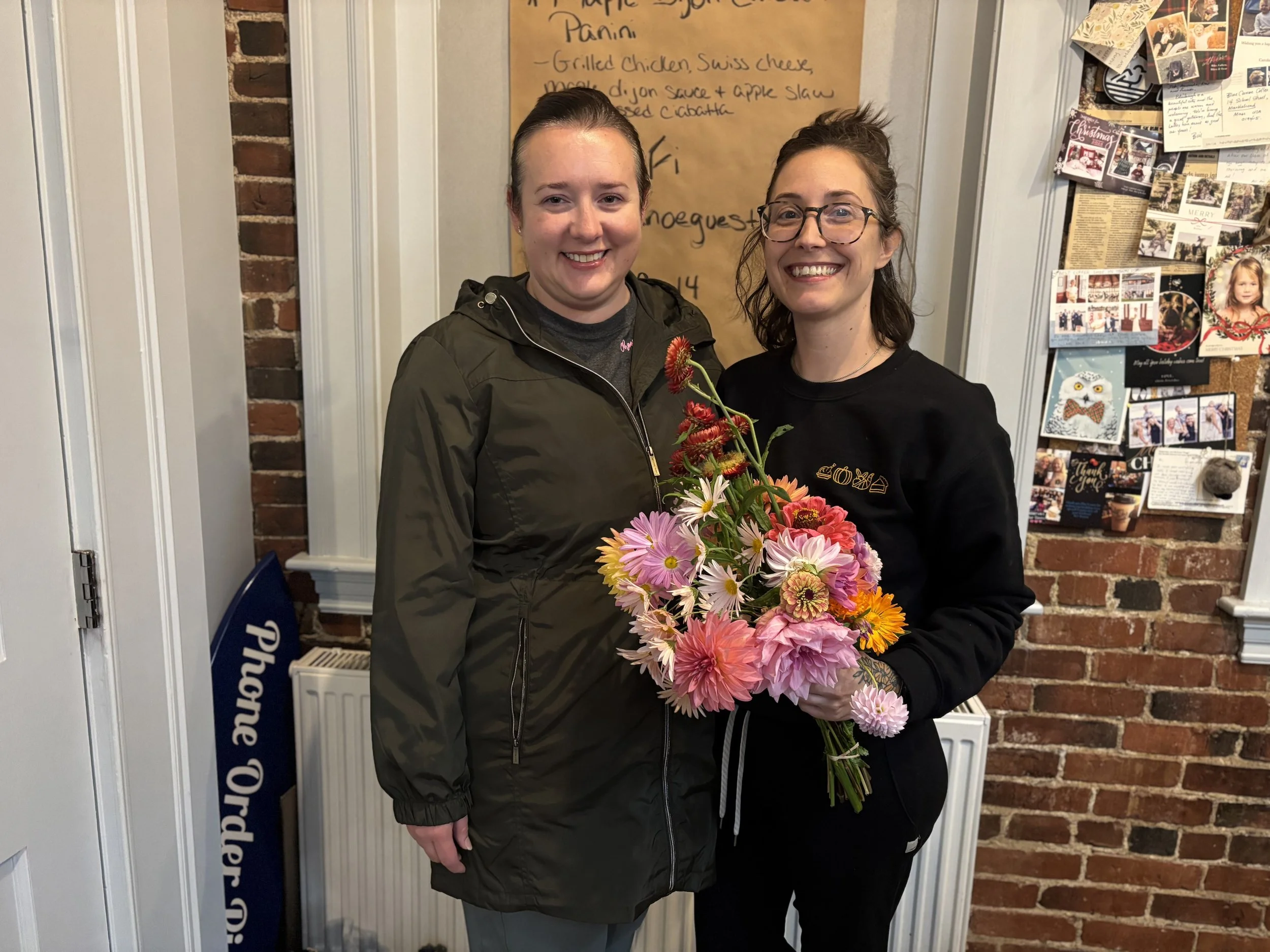 Two women standing indoors, one holding a colorful bouquet of flowers, smiling at the camera.
