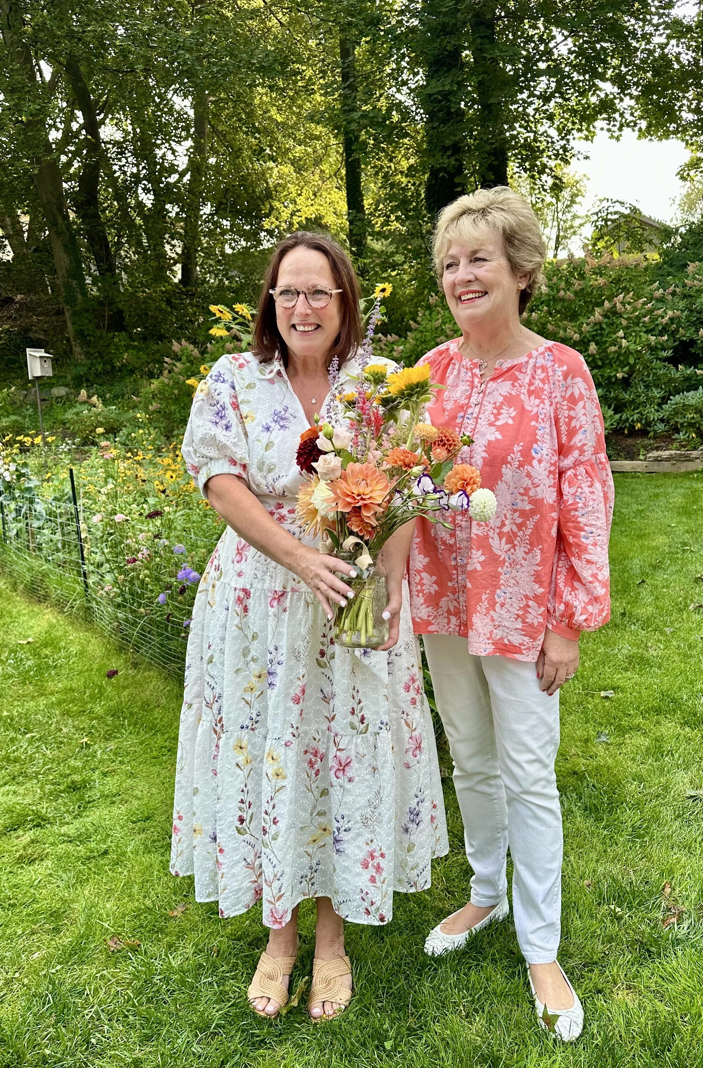 Two women smiling outdoors in a garden, one holding a vase of colorful flowers, both dressed in light, floral clothing.