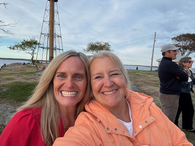 Two women smiling and taking a selfie outdoors by the water, with a tall metal tower, trees, and other people in the background.
