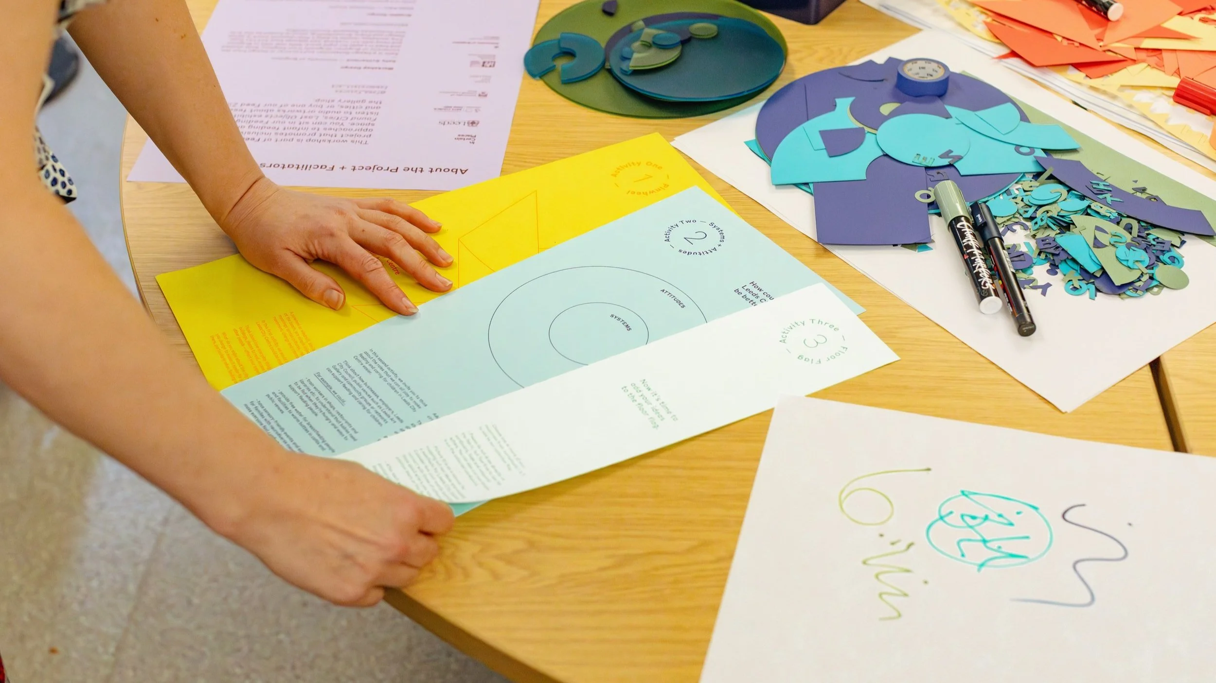 Hands holding three sheets of colourful paper on a table.