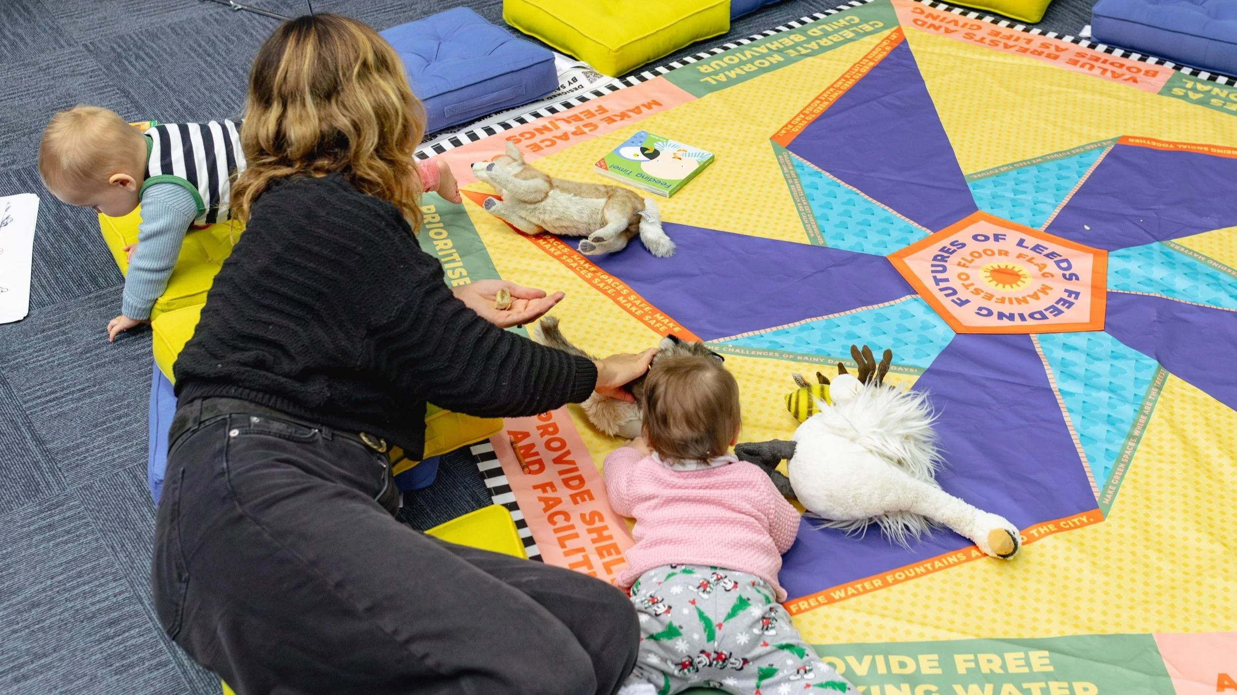 A woman and baby laying on a colourful mat with a pinwheel design in the middle and words, such as 'Provide Shelter and Facilities' around the edges.