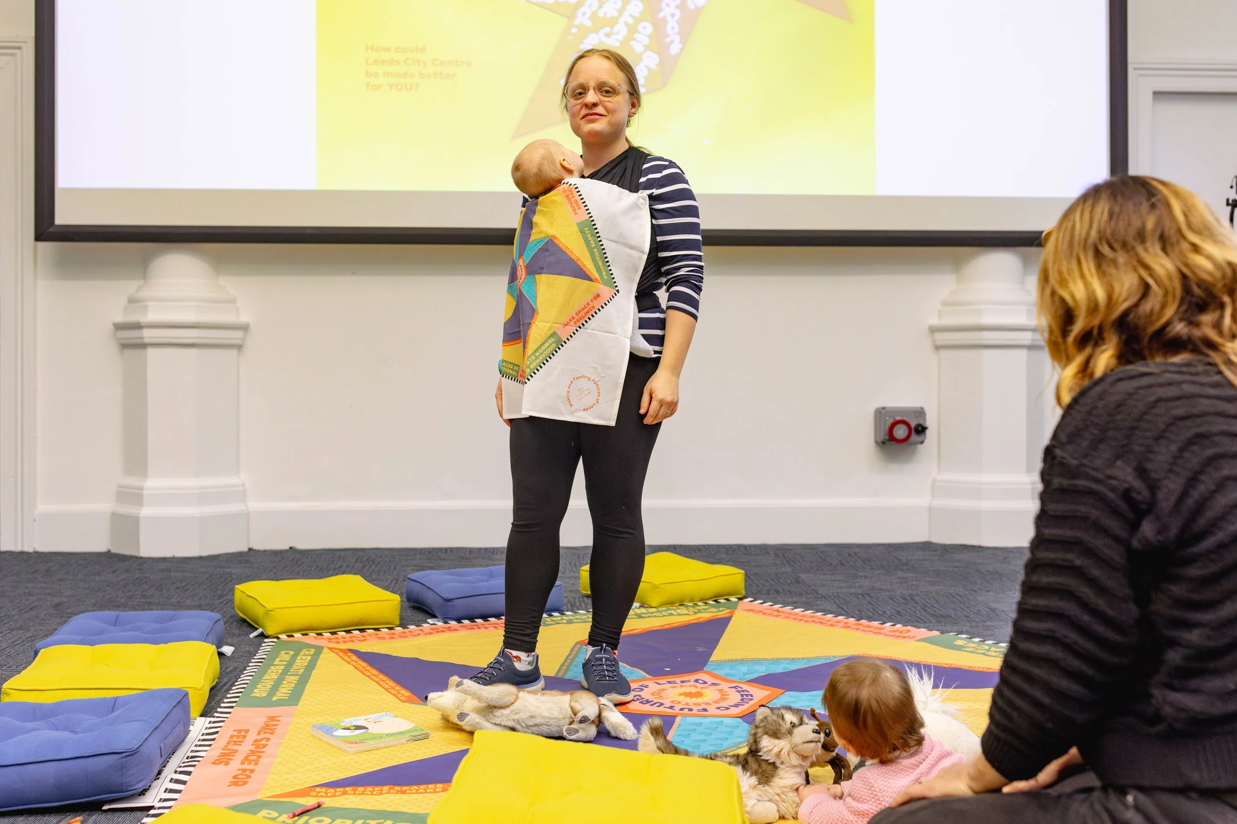 A woman carrying a baby in a sling, with a colourful muslin over the sling, standing on a colourful mat.