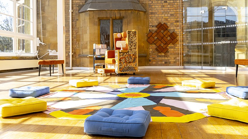 Colourful mat on the floor of a sunny gallery with large windows, surrounded by blue and yellow cushions.