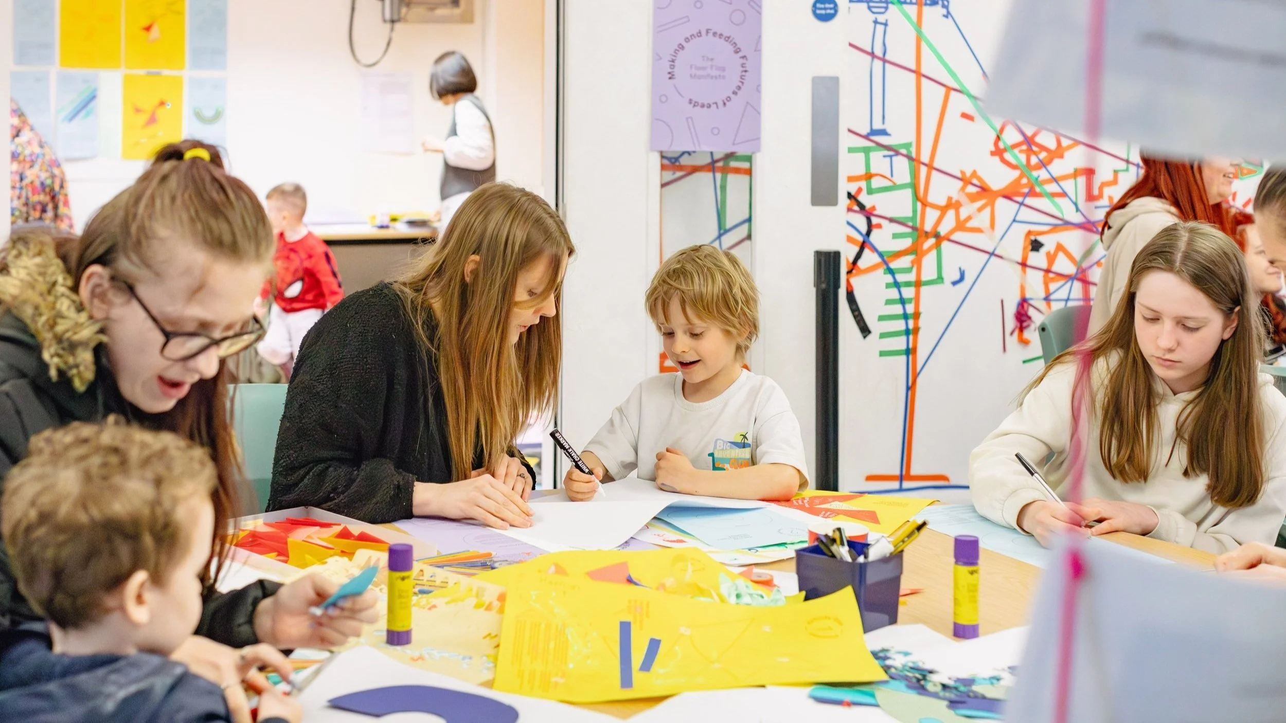 A group of women and children, drawing on colourful worksheets on a table.