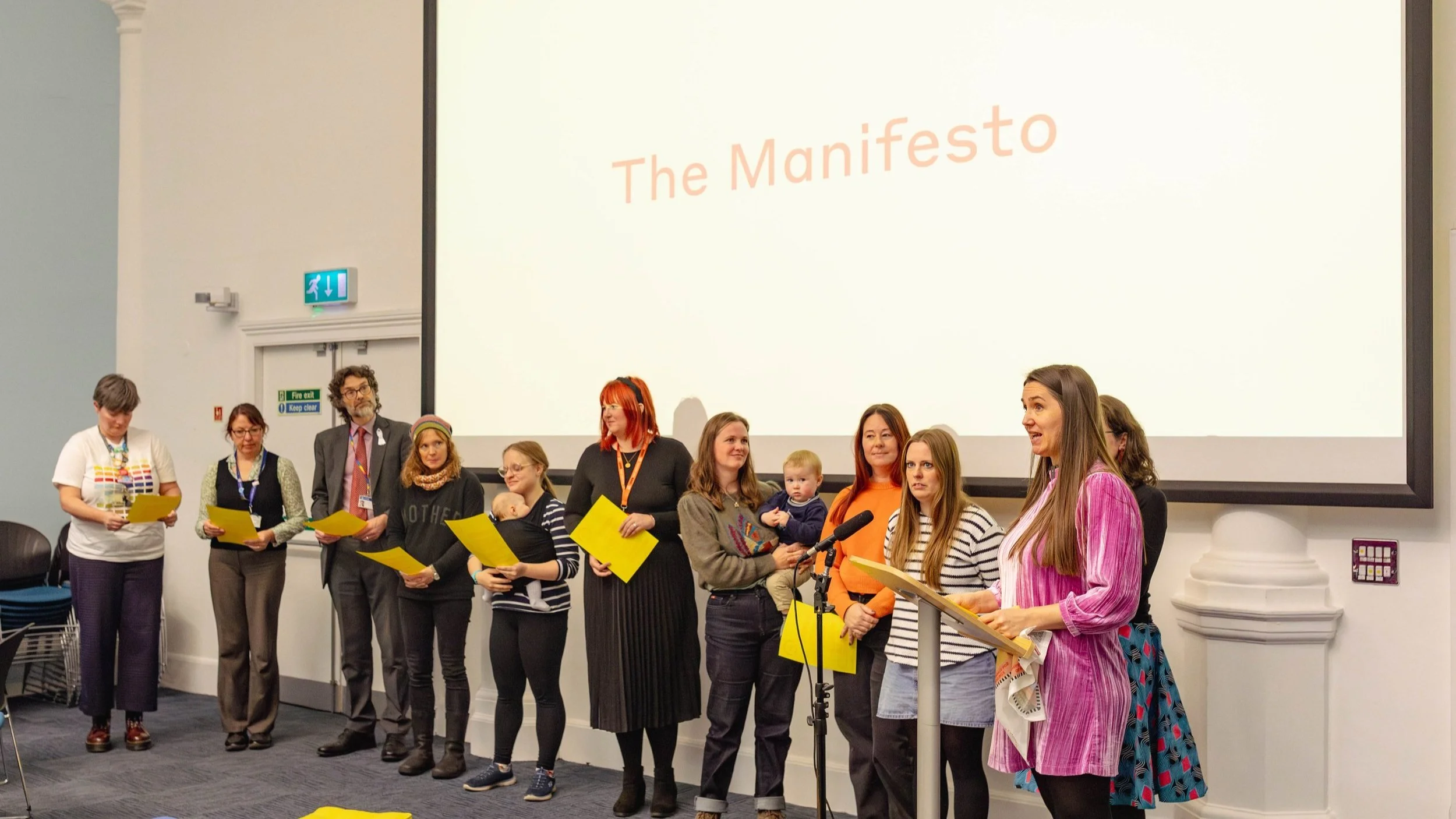 A group of people standing in a line in front of a projection screen with the words 'The Manifesto'