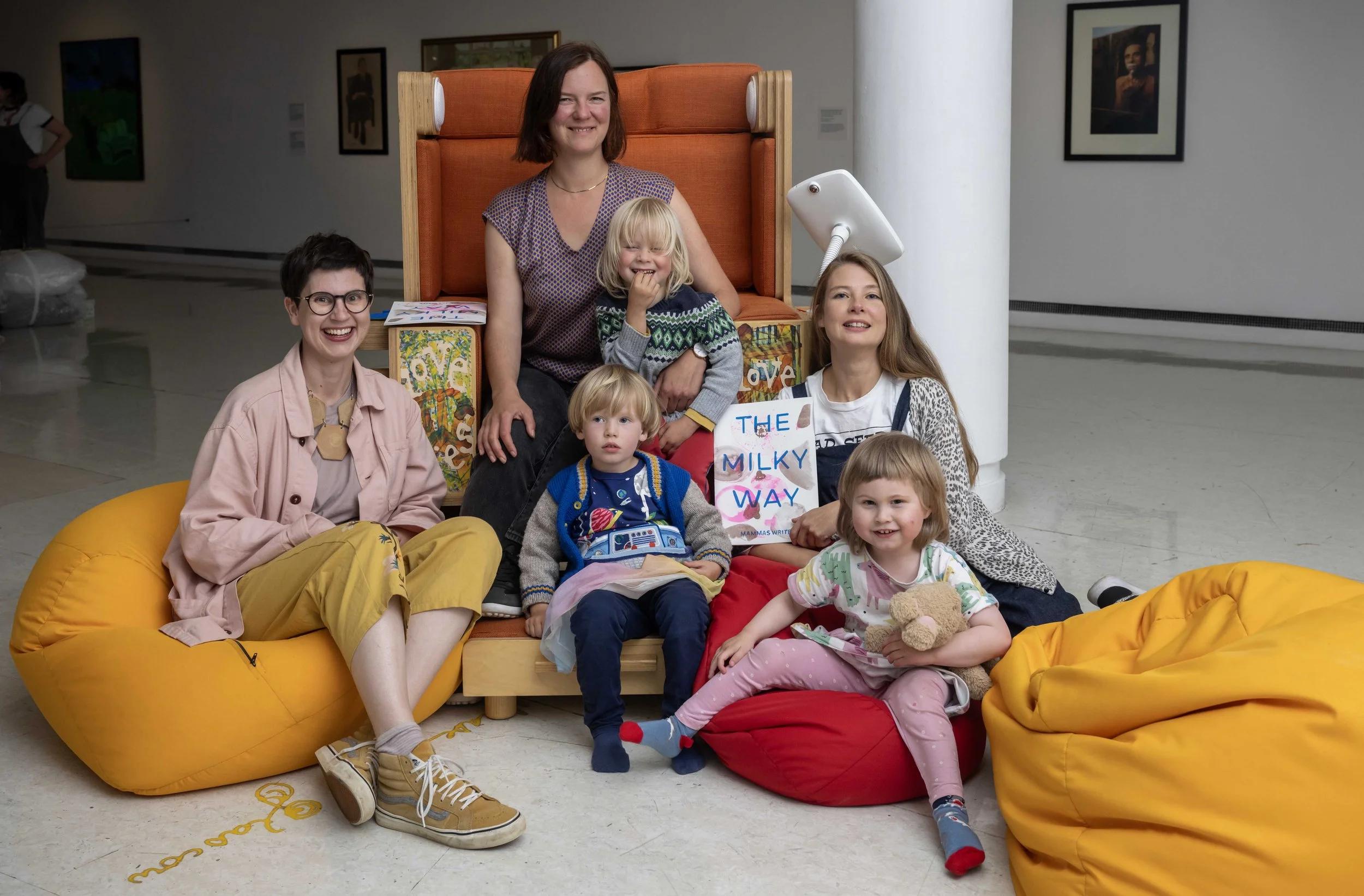 A group of women and children seated around a large orange upholstered chair in a gallery setting