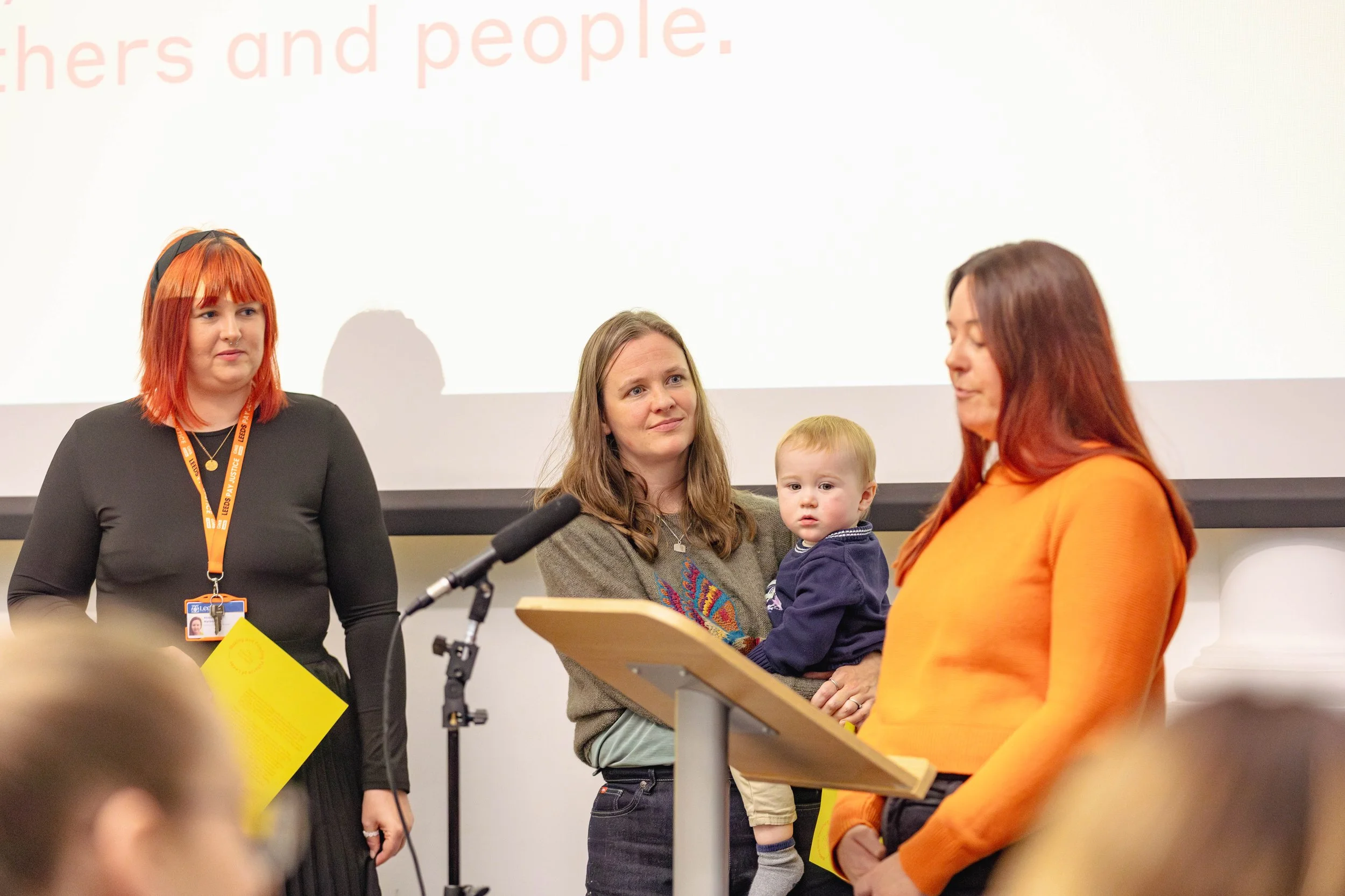 A woman standing at a lectern, surrounded by two other women, one carrying a baby.