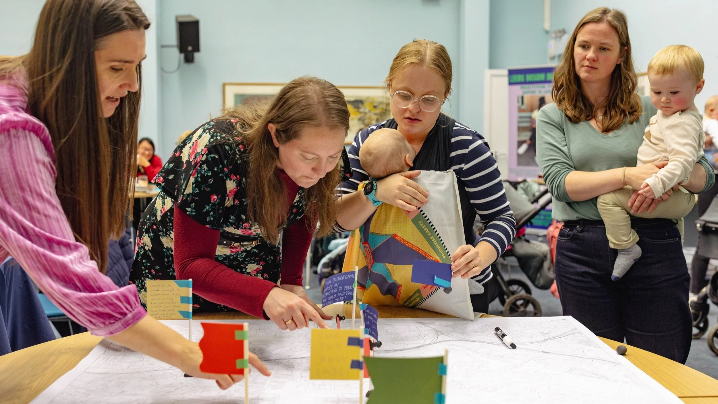 A group of women, one with a baby in a sling, looking at a map on a table top.