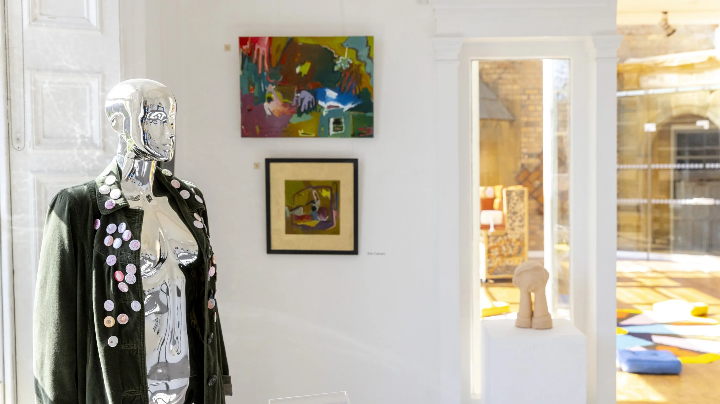 Metallic mannequin wearing a black jacket covered in badges, in front of paintings on a gallery wall.