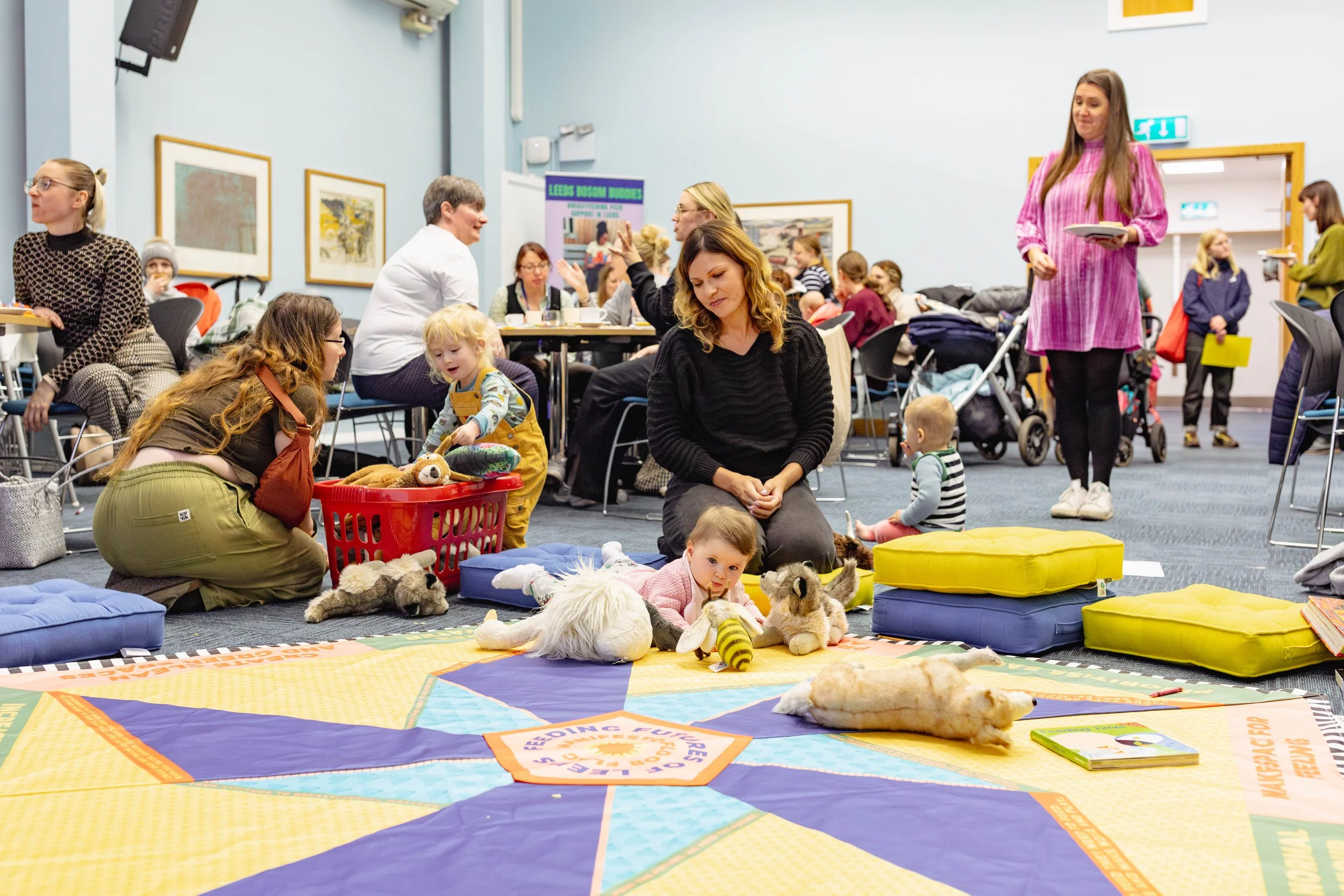 A woman and a baby sitting on a colourful mat in a conference room, surrounded by people sitting on chairs.
