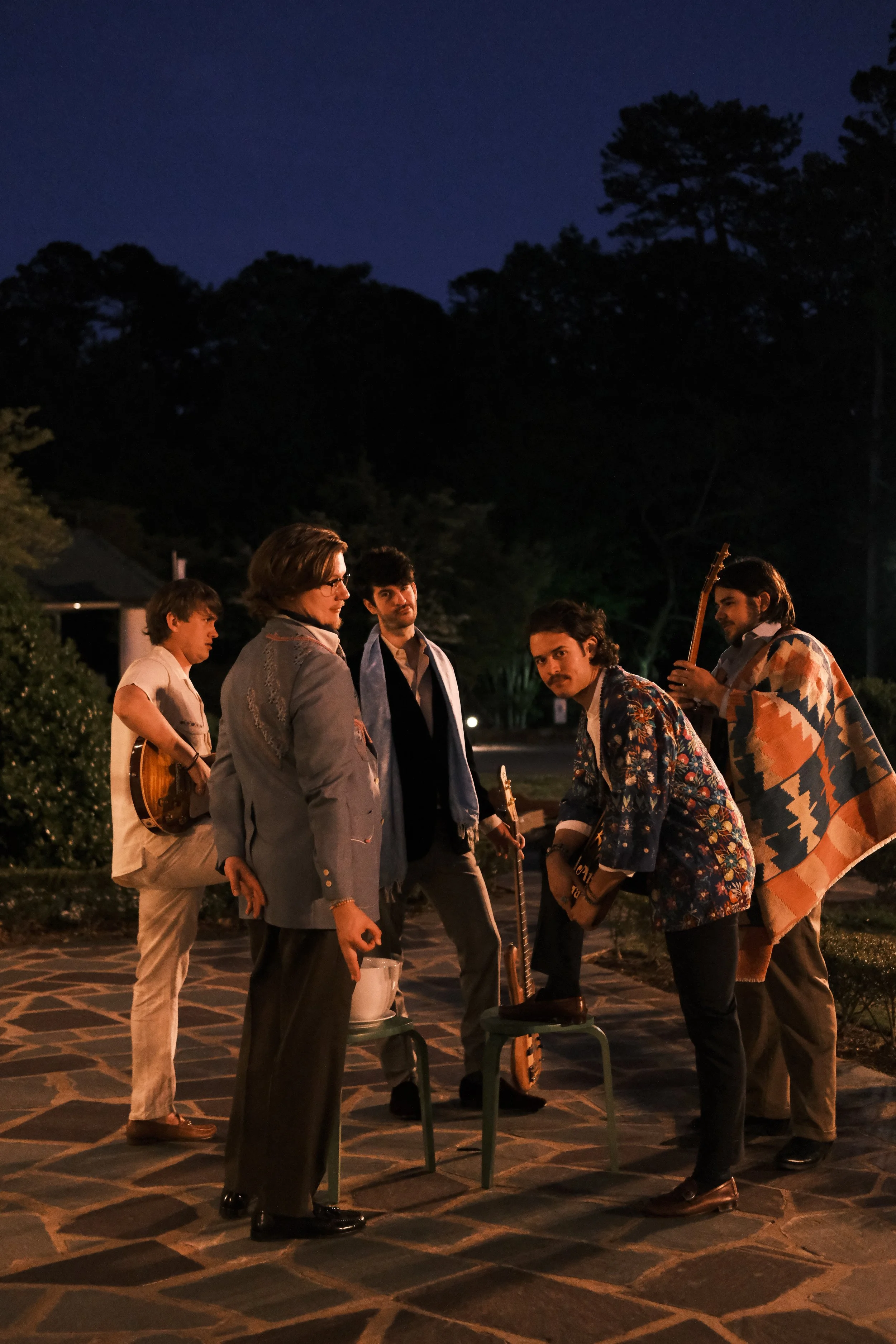 Joe May's Month of Mondays outdoors at night holding guitars, standing on a stone walkway, with two of them wearing patterned jackets and one wrapped in a blanket.