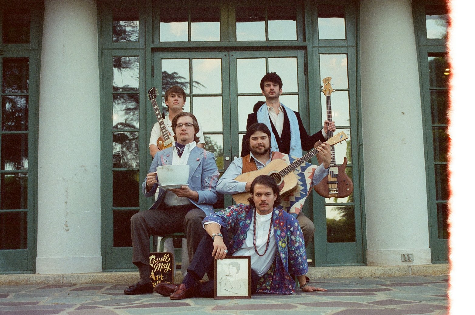 Joe May's Month of Mondays posing with guitars and eccentric objects, in front of a large windowed building.