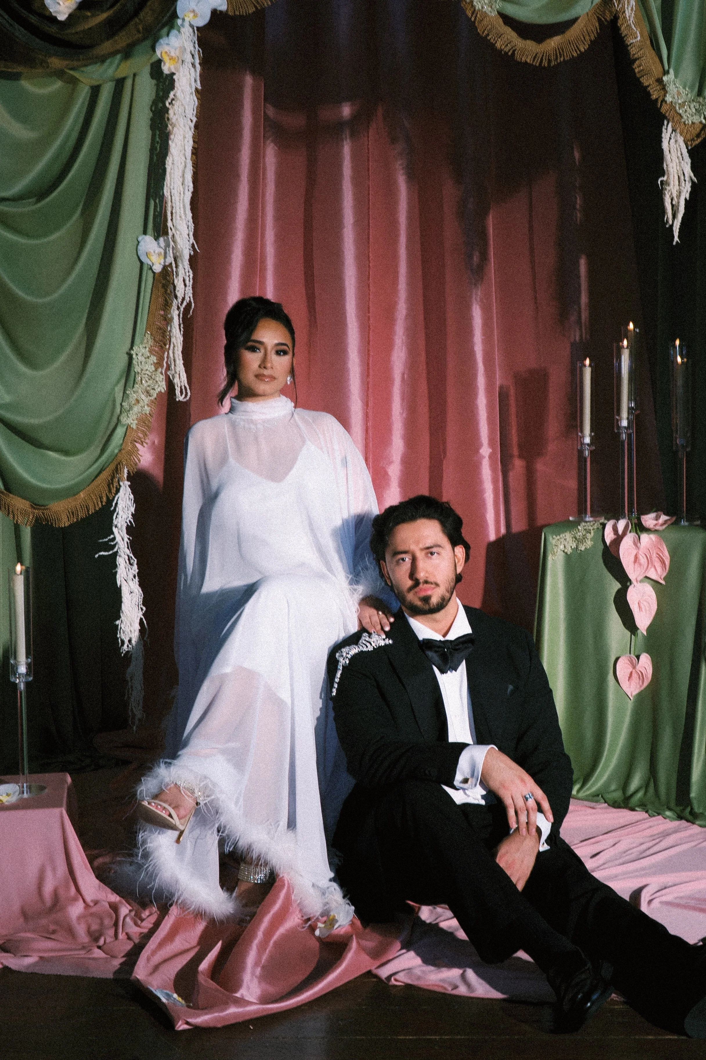 Two quincenera's in princess dresses, one purple and one green, with tiaras and flowers, are posing outdoors among trees and rocks, while two men are photographing them.