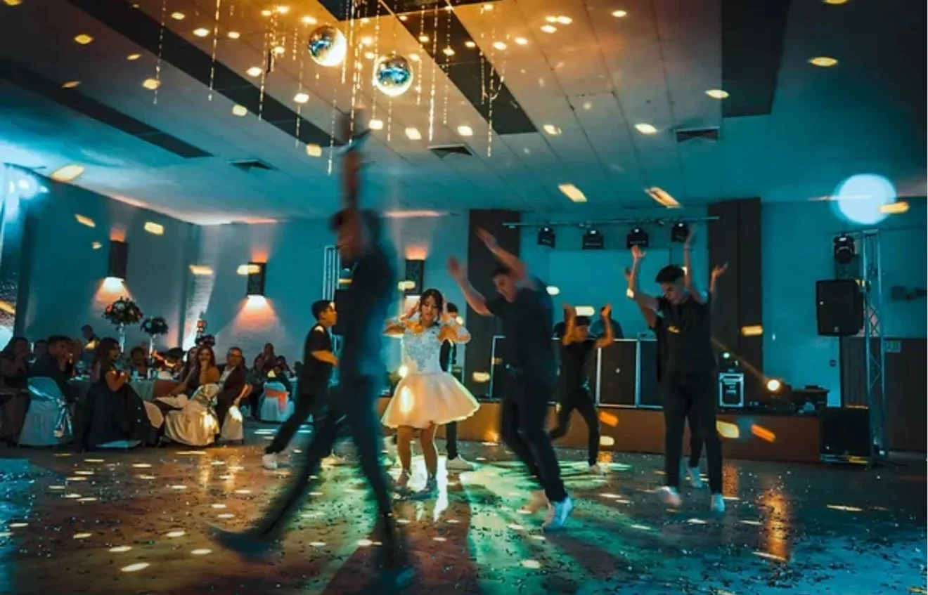 People dancing on a dance floor during a celebration, with guests seated at tables and disco balls hanging from the ceiling.