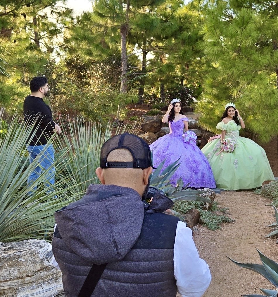 Two quincenera's in colorful dresses and crowns pose in a garden for a photo shoot, with two men nearby and a photographer in the foreground capturing the shot.