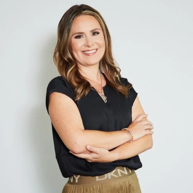 Realtor head shot, smiling woman with long brown hair, wearing a black blouse and a brown pleated skirt, standing against a plain light gray background.
