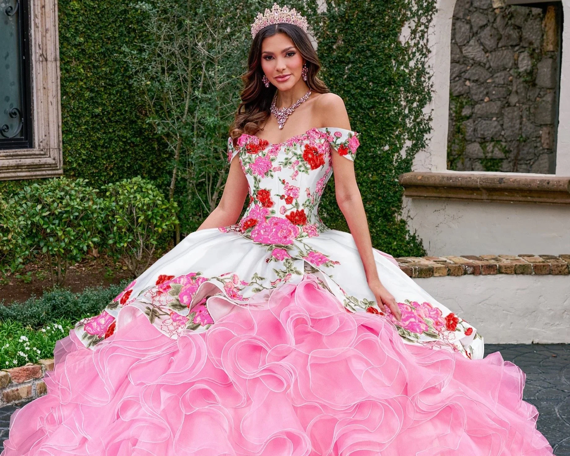 A quincenera smiling woman in a light blue strapless gown with lace details, wearing a tiara, sitting outdoors in front of a terracotta-colored wall and potted plants.